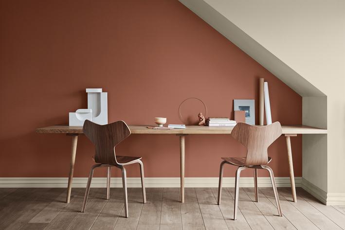 A modern workspace with a wooden desk and two chairs against a terracotta wall, beneath an angled cream ceiling.