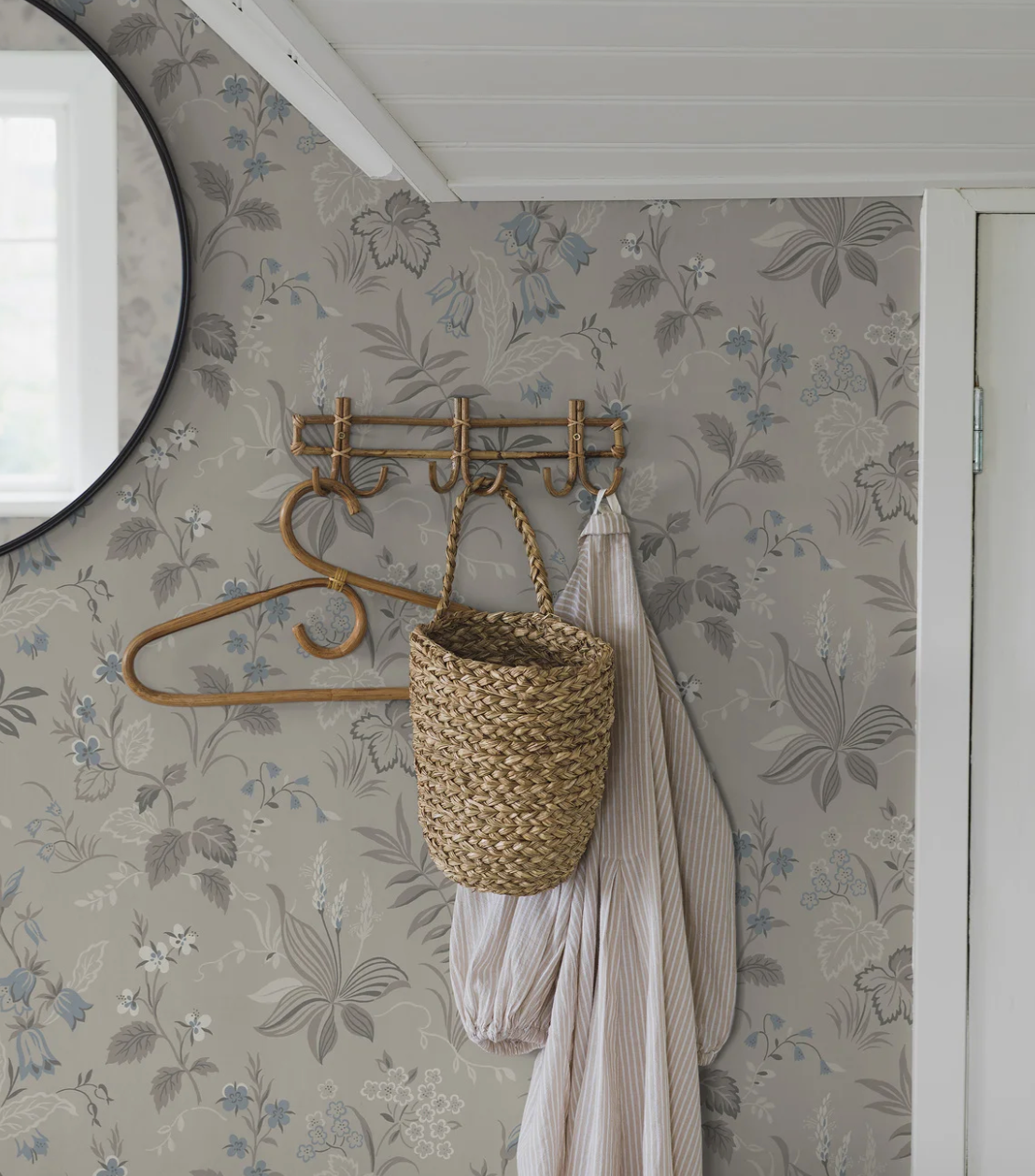 A wall with gray floral wallpaper, a rattan coat rack holding a woven basket and striped shirt, and a round mirror.