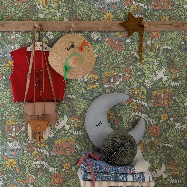 A wall with green wallpaper showing houses, trees, and birds. A red vest, straw hat, and star wand hang from a wooden peg rail. A moon pillow rests on folded textiles below.