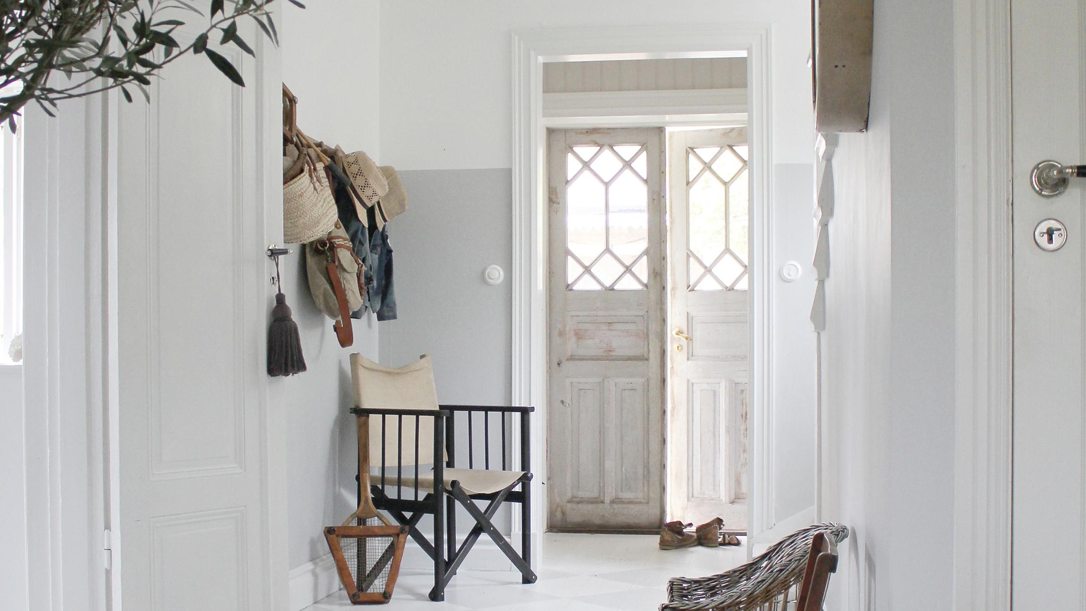 A bright hallway with white and gray walls, featuring a coat rack, a director's chair, and an open doorway to a rustic double door.