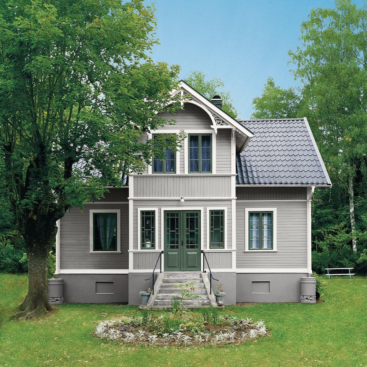 A light gray house with dark green doors and window frames, a dark tiled roof, set in a green lawn with trees.