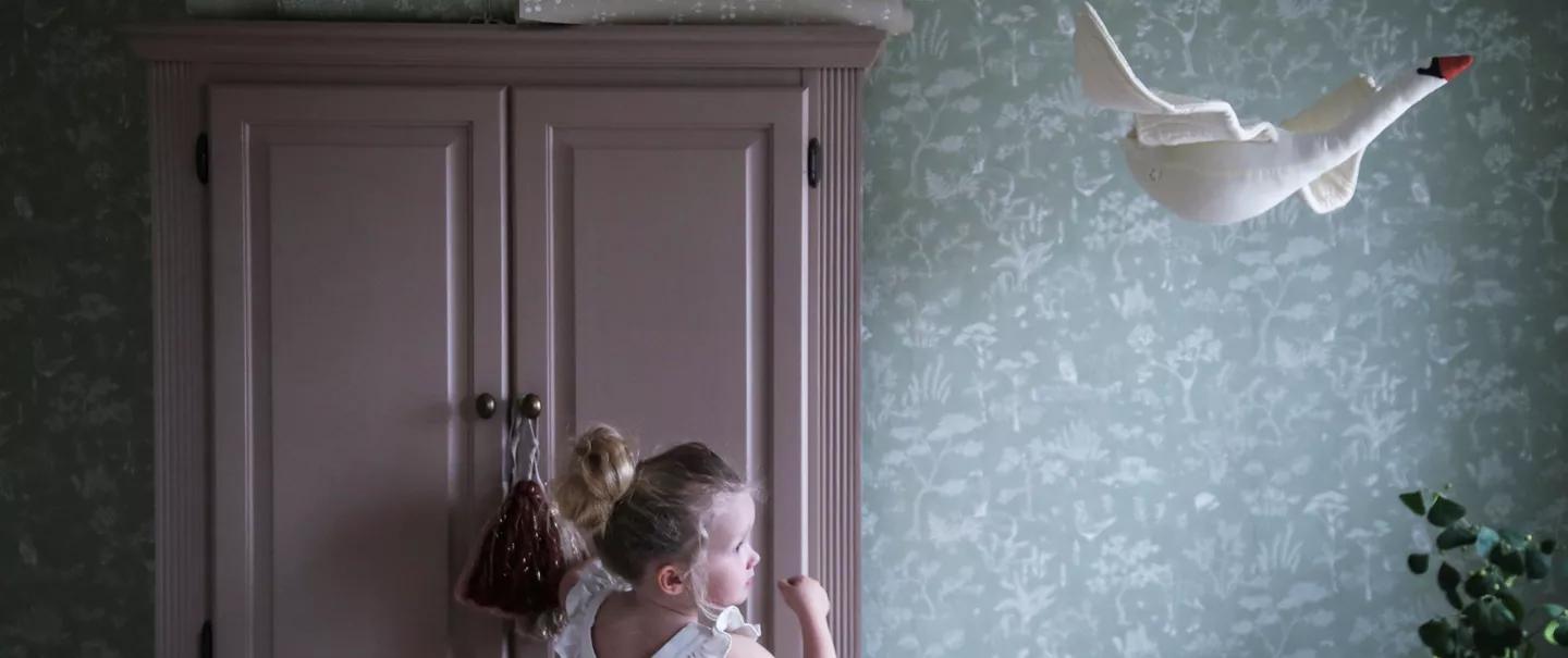 A young girl looks up at a white swan hanging on a patterned green wall, with a pink cabinet behind her.
