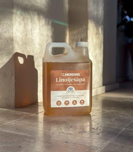 A clear plastic bottle of Herdins Linoljesåpa (linseed oil soap) with amber liquid, sitting on a tiled floor with strong sunlight casting shadows.