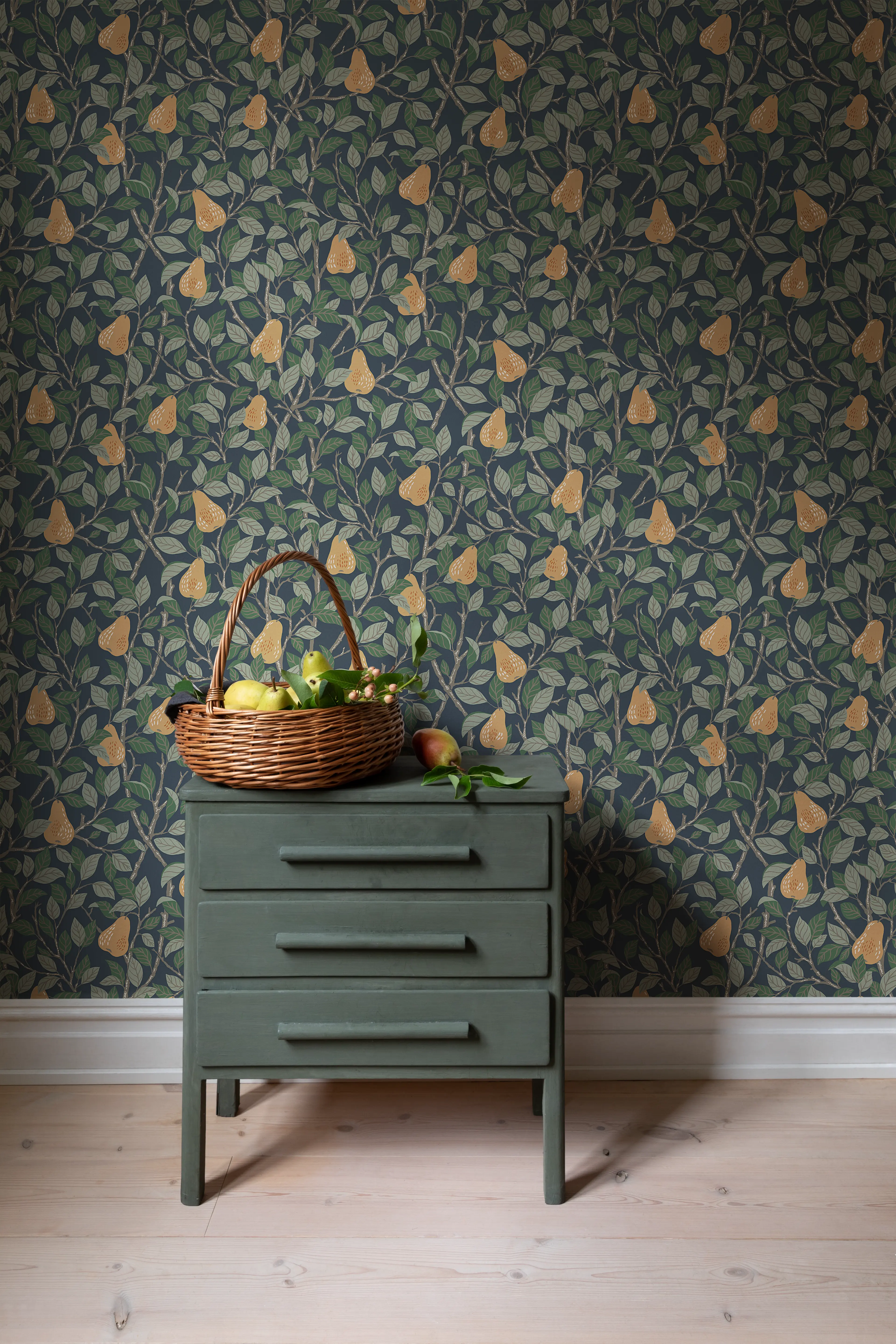Dark blue wallpaper with a pattern of green leaves and yellow pears, behind a dark green chest of drawers topped with a fruit basket.
