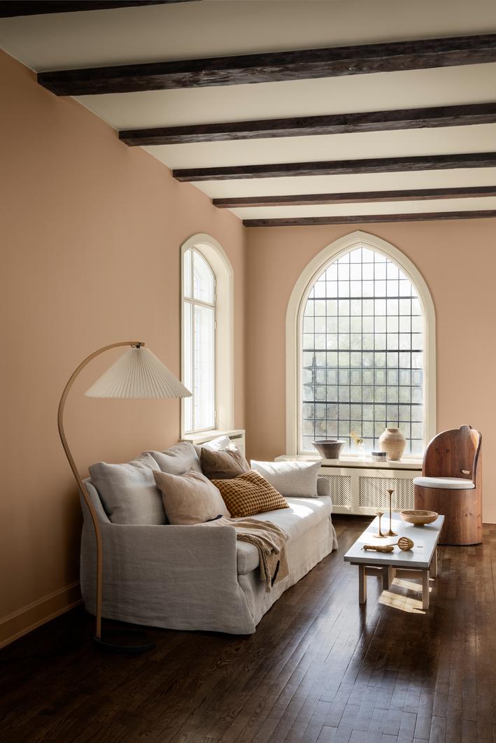 A living room with tan walls, dark exposed ceiling beams, arched windows, a light gray sofa, a floor lamp, and a wooden accent chair.