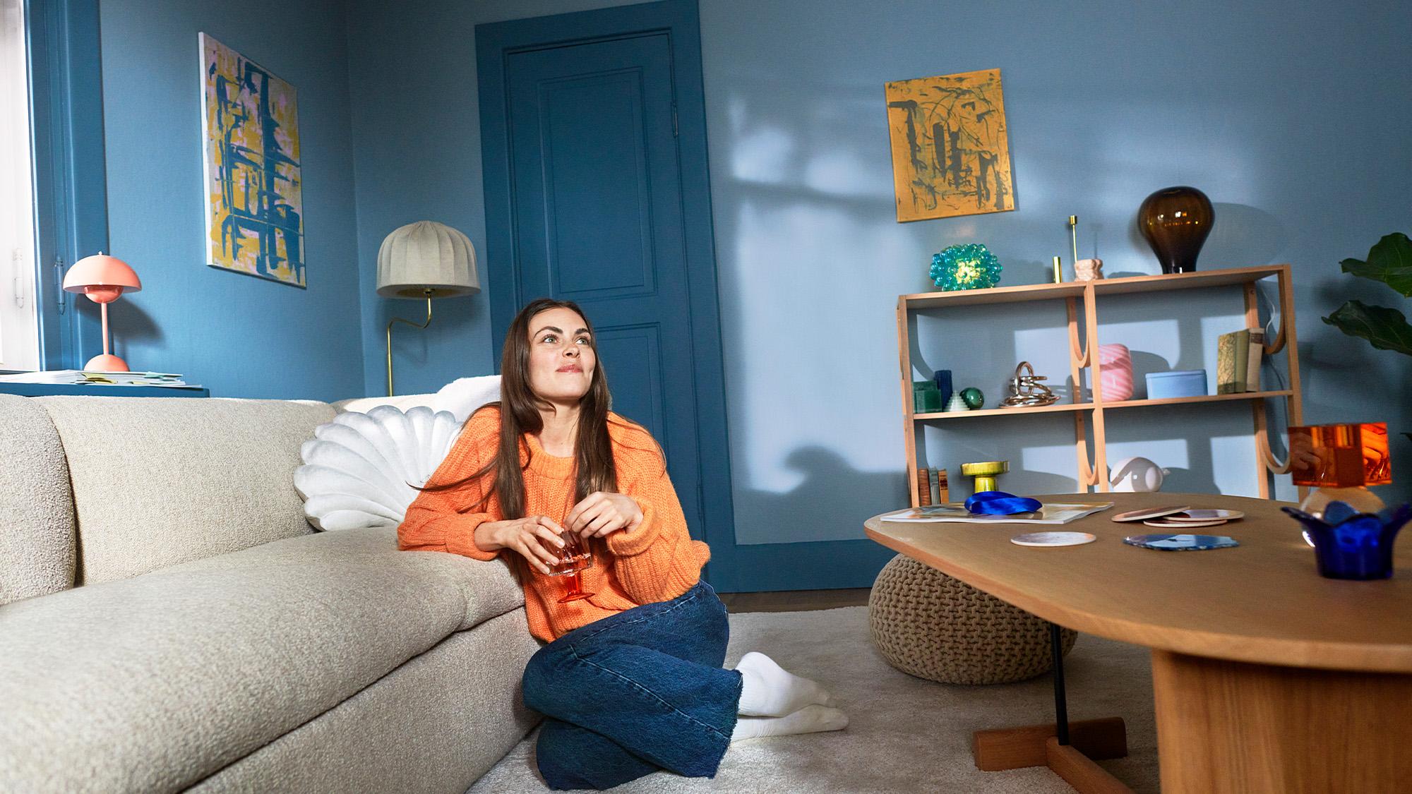 A woman sits on the floor leaning against a sofa in a colorful living room, holding a drink.