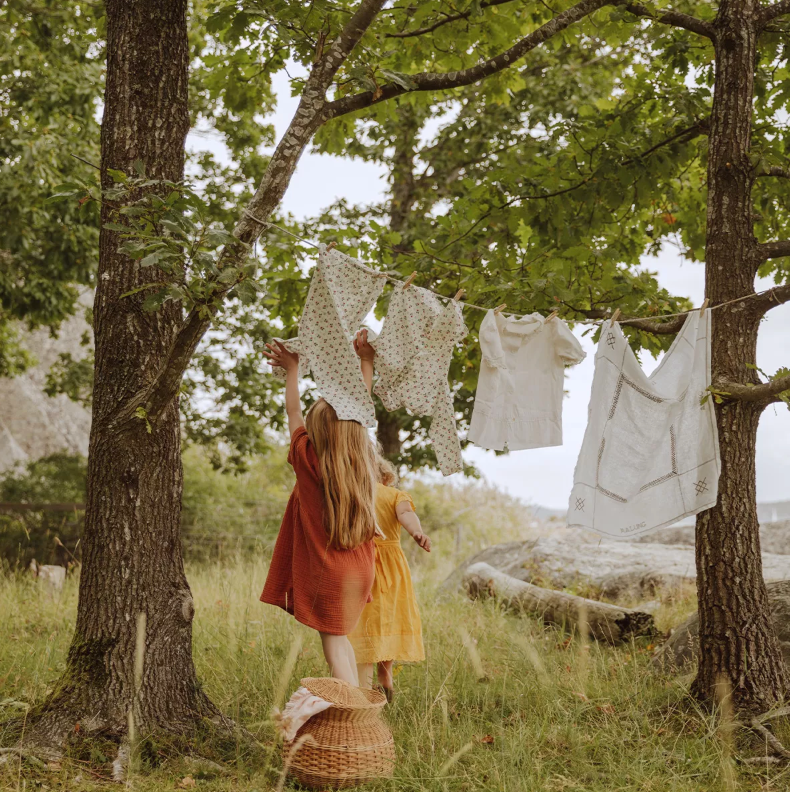 Two young girls, seen from behind, hang laundry on a clothesline strung between trees in a grassy setting.