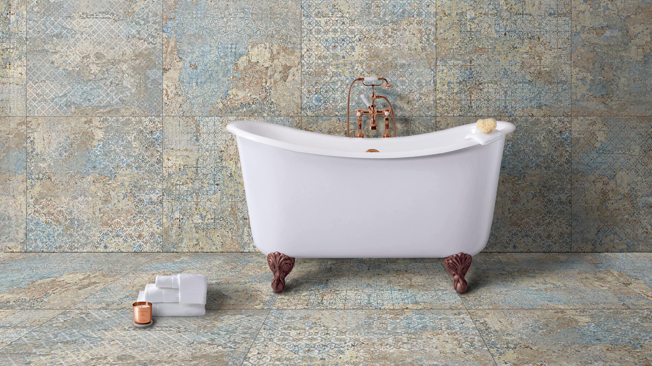 White clawfoot tub with copper fixtures in a bathroom featuring distressed blue and beige patterned tiles, with towels and a candle on the floor.