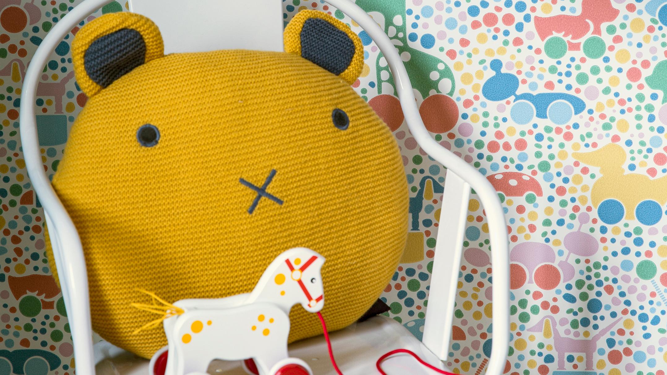 A yellow bear pillow and a toy horse in a white chair, against colorful toy-patterned wallpaper.