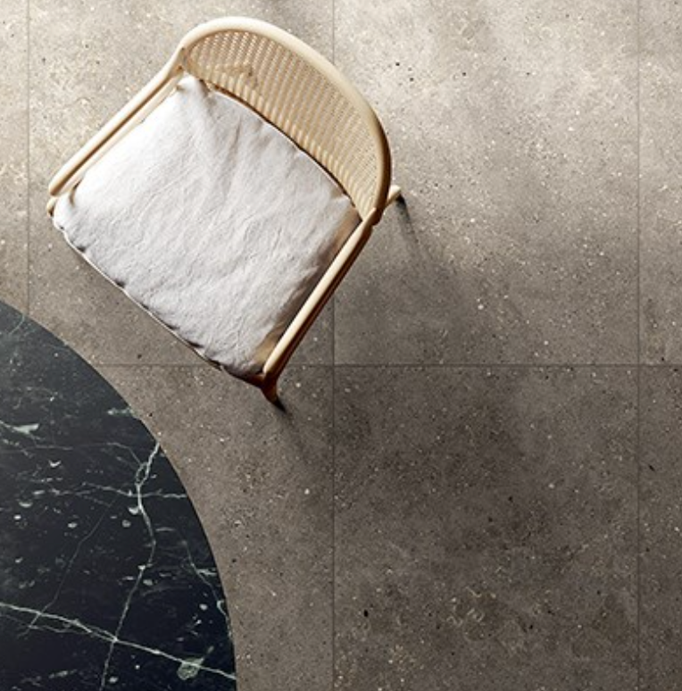 Overhead view of a light woven chair with a cushion on dark grey speckled tiles, next to a curved dark marble surface.