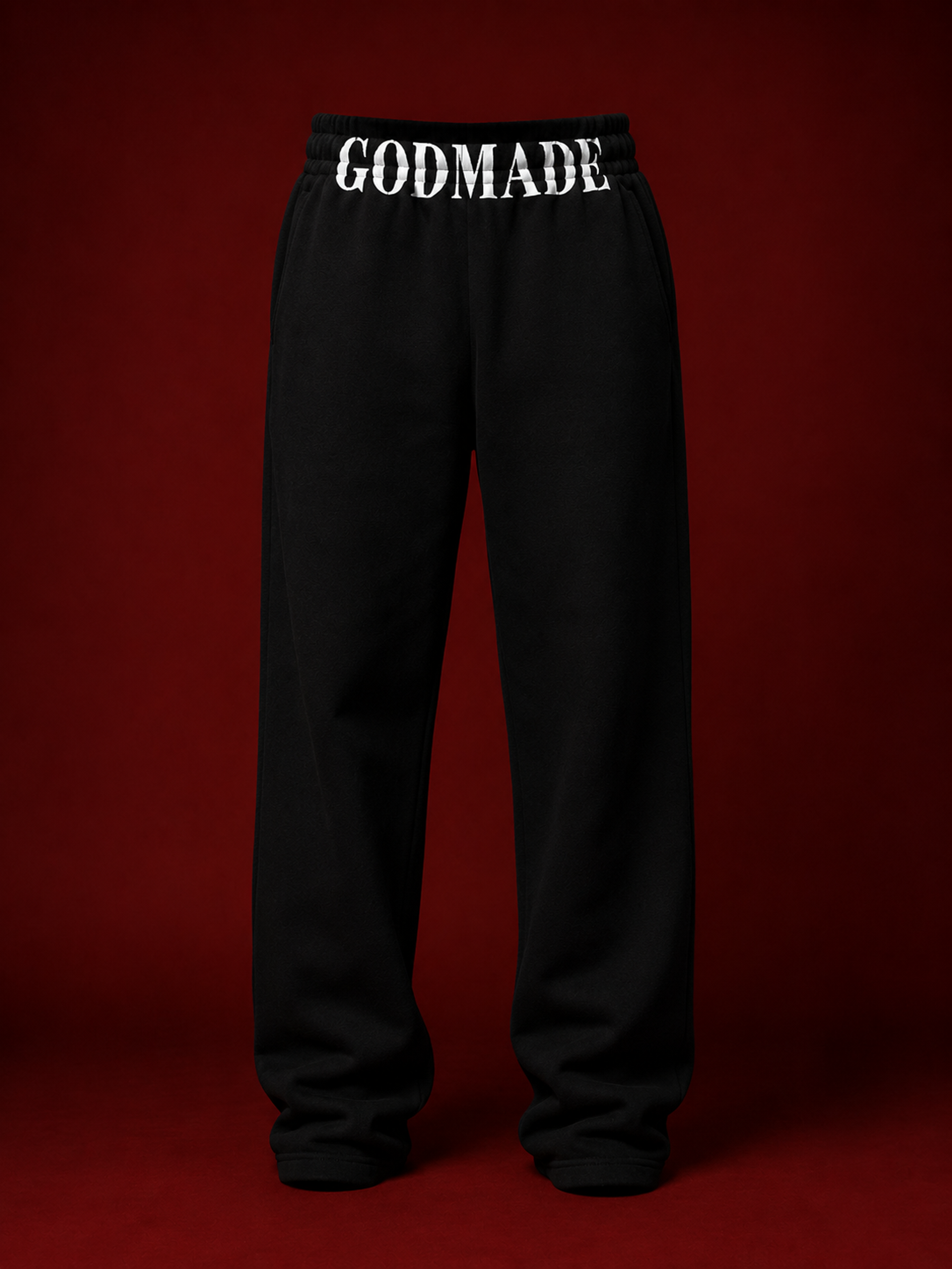 Black oversized GODMADE sweatpants with bold white GODMADE lettering across the waistband, displayed in a luxury studio setup against a deep burgundy background.