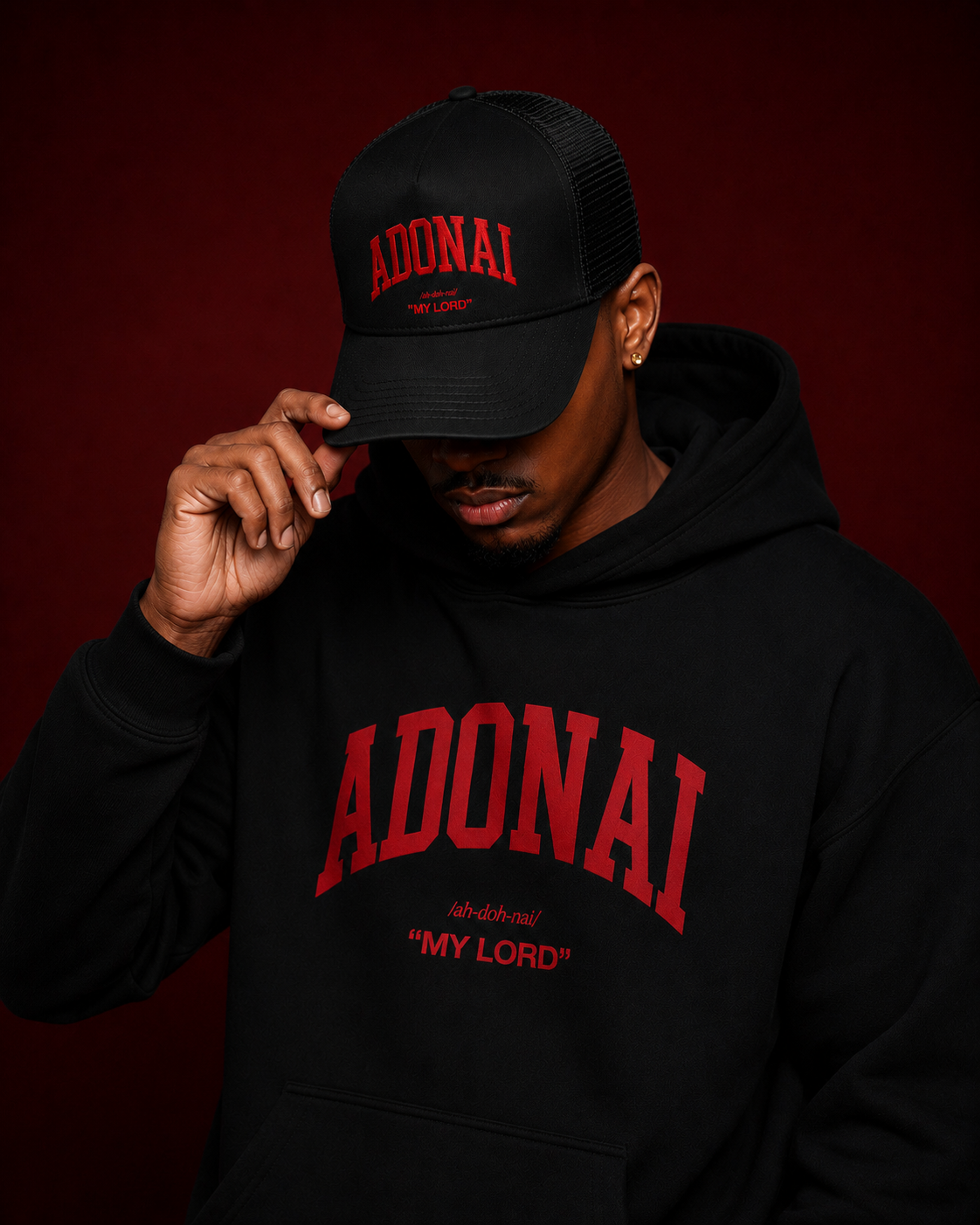Black trucker cap featuring bold red embroidered “ADONAI” lettering with “My Lord” beneath it, photographed against a rich burgundy background with dramatic studio lighting.