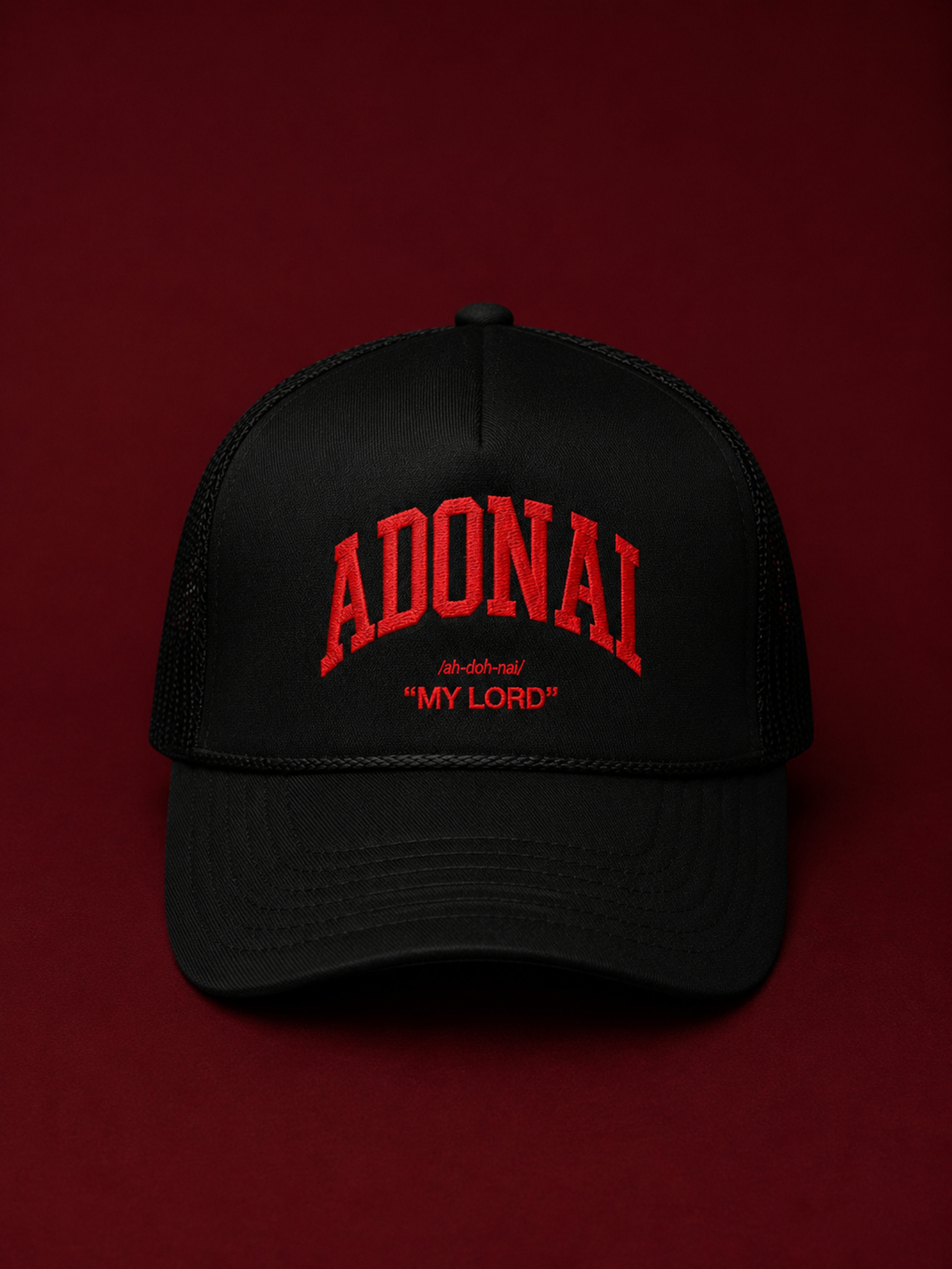 Black trucker cap featuring bold red embroidered “ADONAI” lettering with “My Lord” beneath it, photographed against a rich burgundy background with dramatic studio lighting.