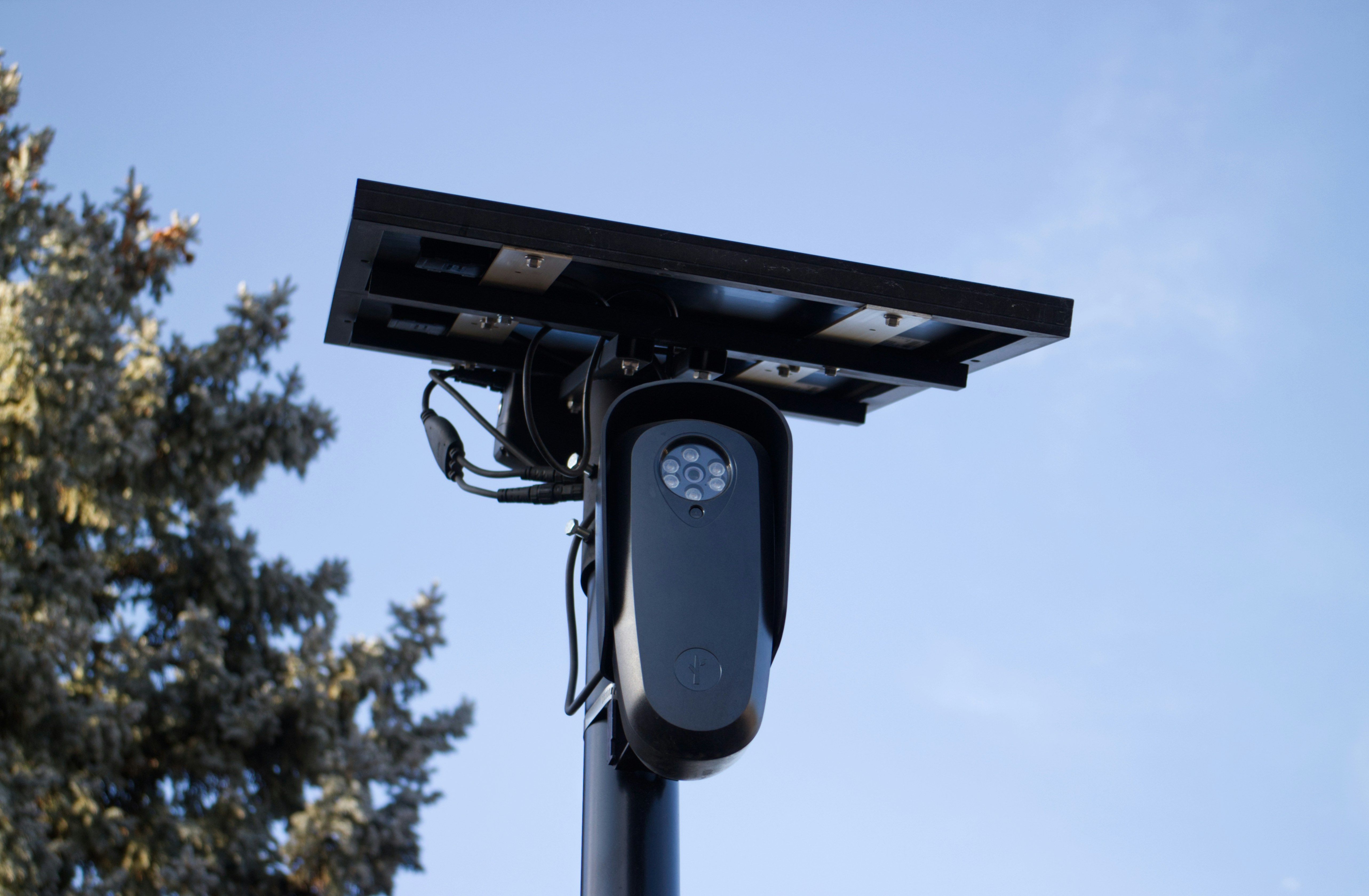 License plate reader surveillance camera with a solar panel mounted on a pole against a blue sky.