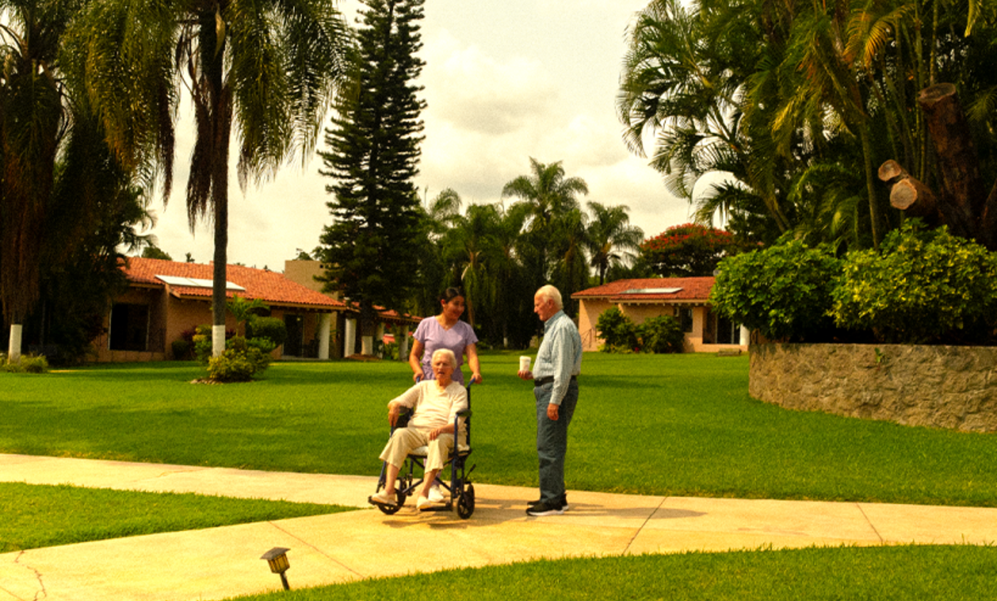 Residencia de retiro con exclusividad, entorno verde, sereno, tranquilo, instalaciones dignas, enfermera pasea a un residente en silla de ruedas en compañía de un amigo
