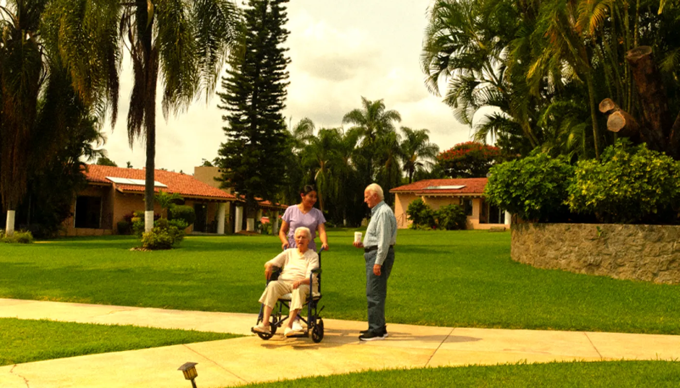 Residencia de retiro con exclusividad, entorno verde, sereno, tranquilo, instalaciones dignas, enfermera pasea a un residente en silla de ruedas en compañía de un amigo