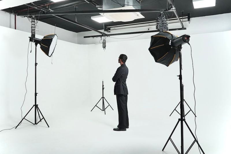Man in formal suit standing in a professional white cyclorama studio setup with softbox lights, inspecting lighting arrangement for a commercial photoshoot or video production