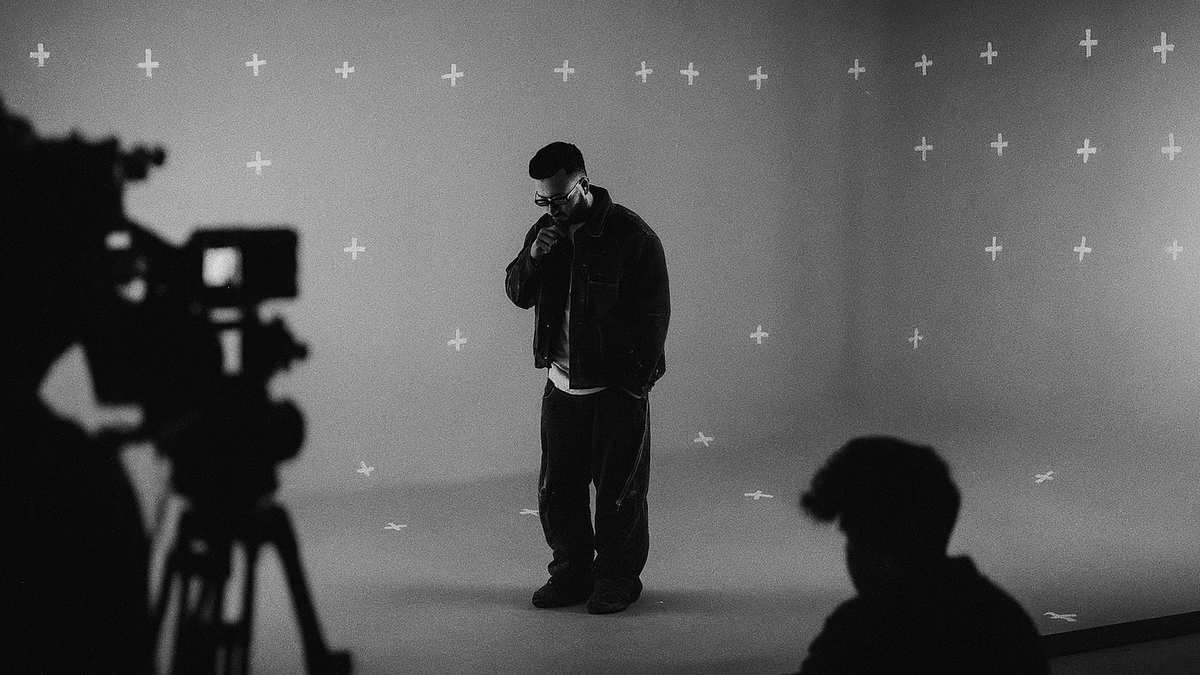 Black and white wide-angle photo of a cinematic studio shoot with a male artist standing under soft lighting, surrounded by camera crew silhouettes and VFX tracking markers on the background wall, capturing a raw behind-the-scenes moment in a professional production setup