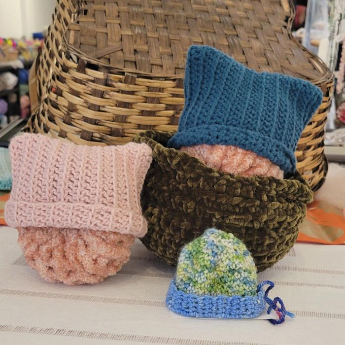 A photo of 3 knit baby hats in front of a basket. The hat colors are pink, blue and green.