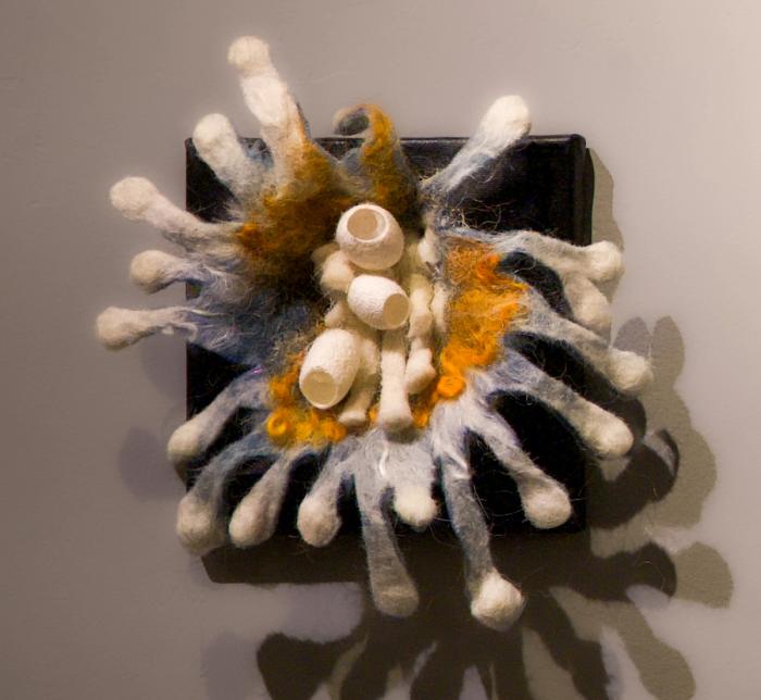 Photo of a smallfelted 3-D organic shaped sculpture. Colors are grey, white, orange and black.