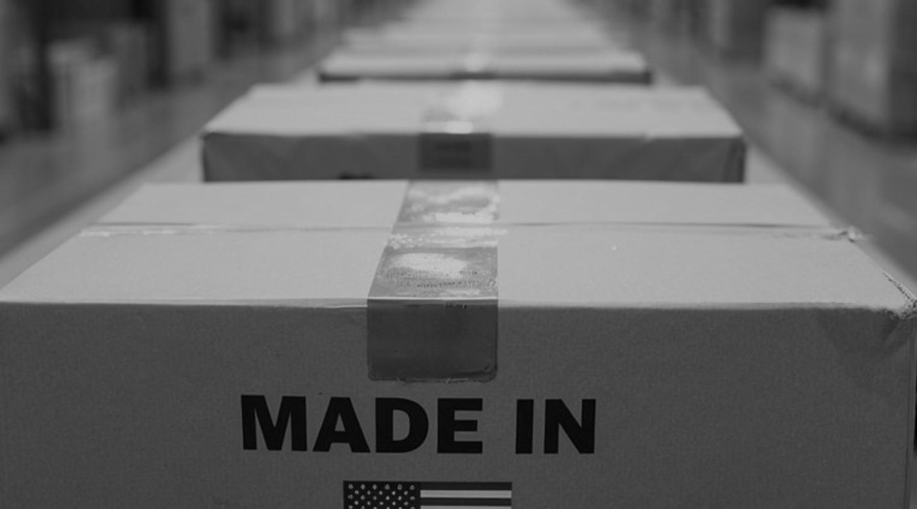 Photograph of sealed cardboard shipping boxes moving along a warehouse conveyor system, with the nearest box labeled “Made in USA” and displaying a U.S. flag. The background shows a blurred industrial warehouse setting with shelving and overhead lighting.