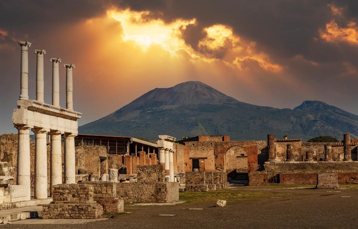 the sun is setting over the ruins of pompeii with a volcano in the background .