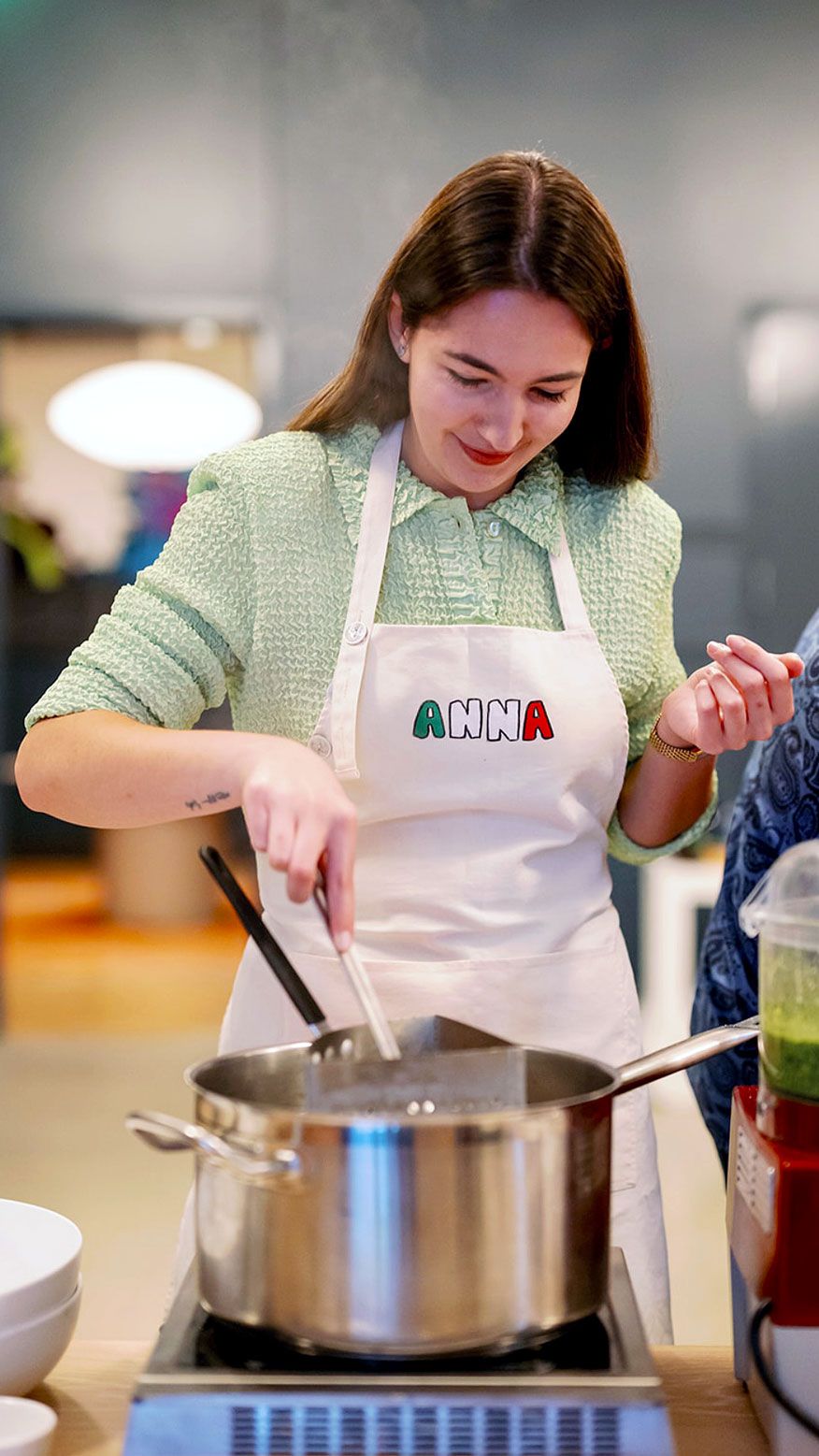 A smiling woman named Anna stirs a steaming pot on a stovetop.