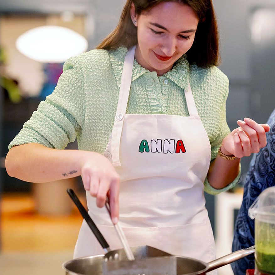 A smiling woman named Anna stirs a steaming pot on a stovetop.