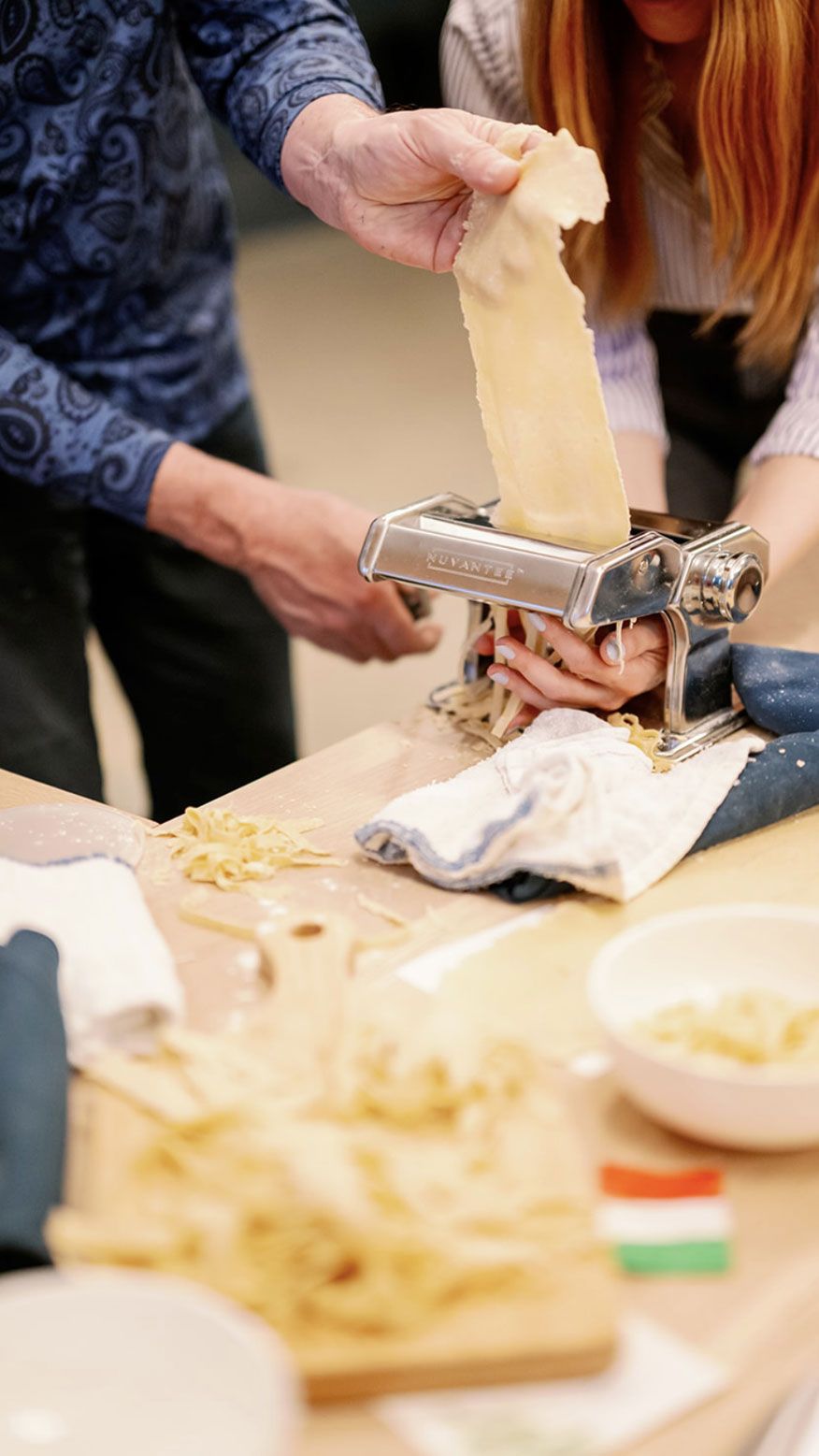 Hands feed pasta dough into a silver Nuvantie machine, slicing it into fettuccine. Fresh pasta and a small Italian flag are on the wooden counter.