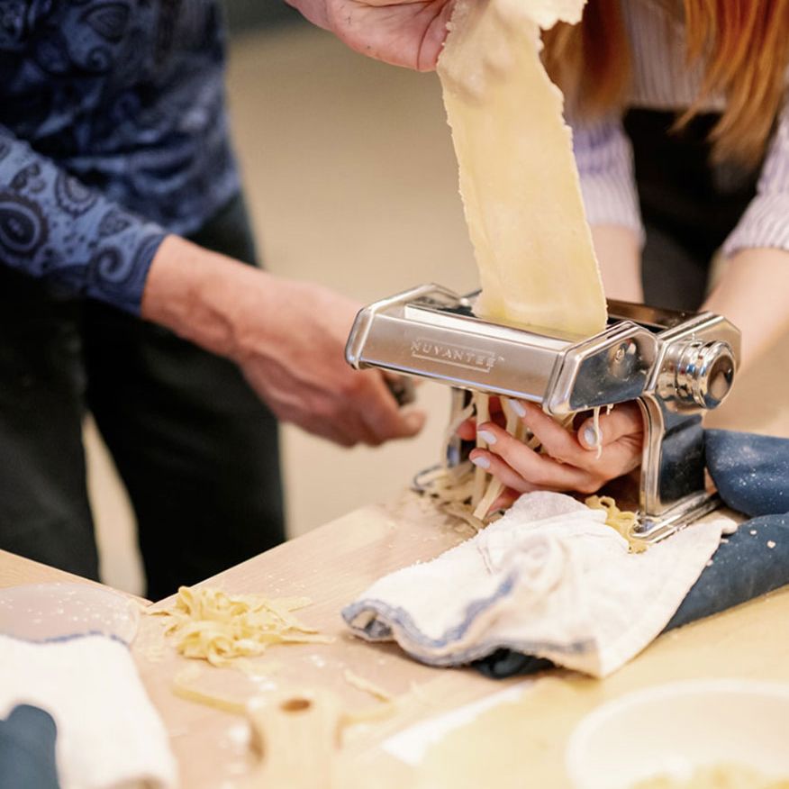 Hands feed pasta dough into a silver Nuvantie machine, slicing it into fettuccine. Fresh pasta and a small Italian flag are on the wooden counter.