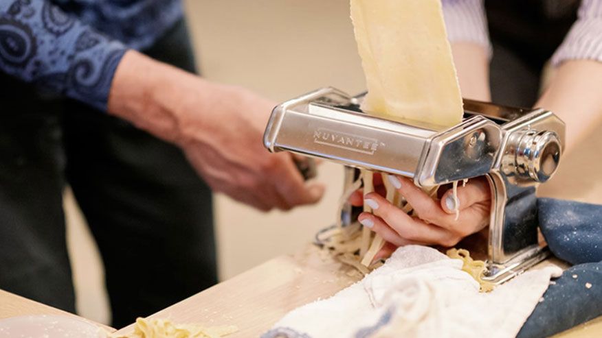 Two people feed pasta dough into a manual pasta maker, creating fresh noodles on a wooden table with an Italian flag nearby.
