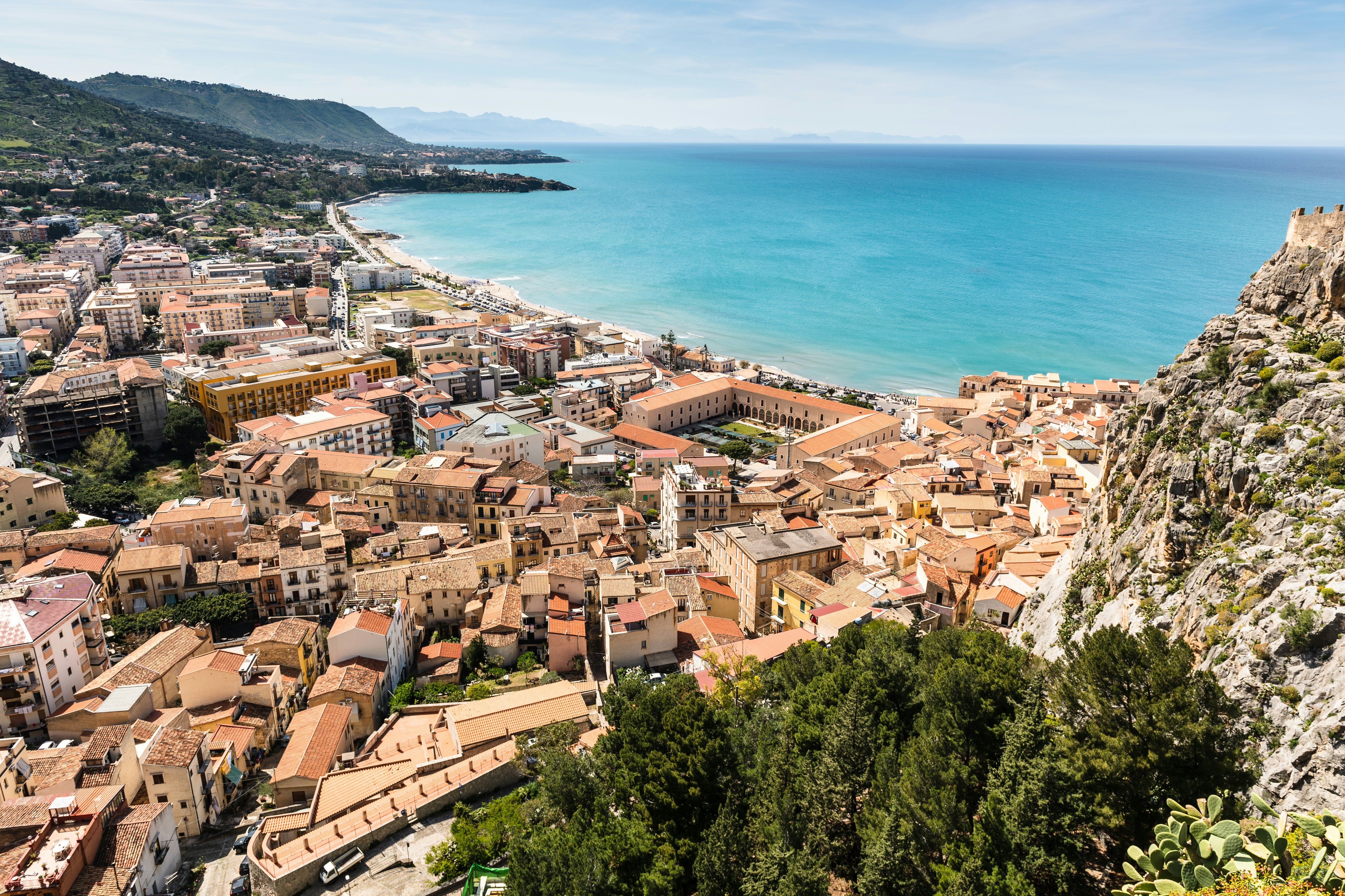 A high-angle view of a coastal town with terracotta-roofed buildings beside a sandy beach and bright blue sea, with a large rocky cliff on the right.