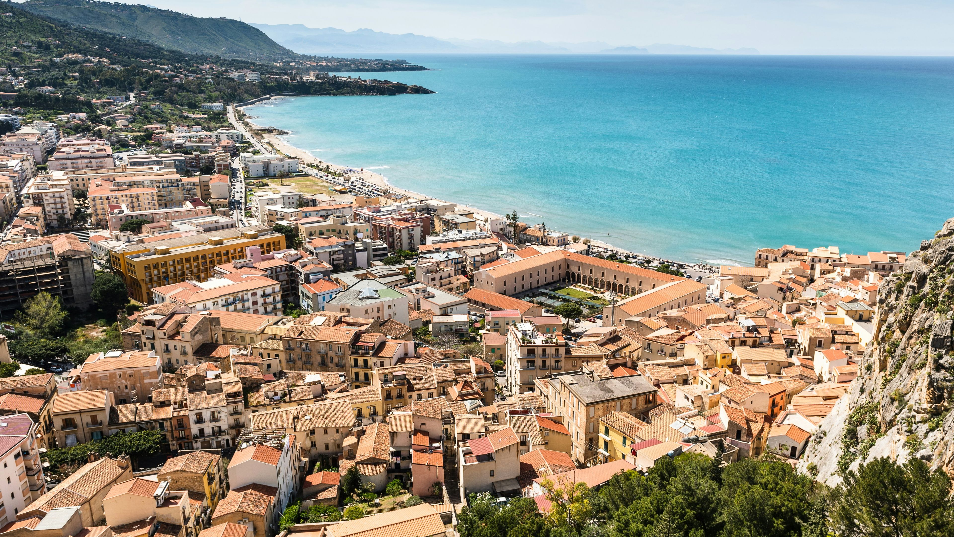 A high-angle view of a coastal town with terracotta-roofed buildings beside a sandy beach and bright blue sea, with a large rocky cliff on the right.