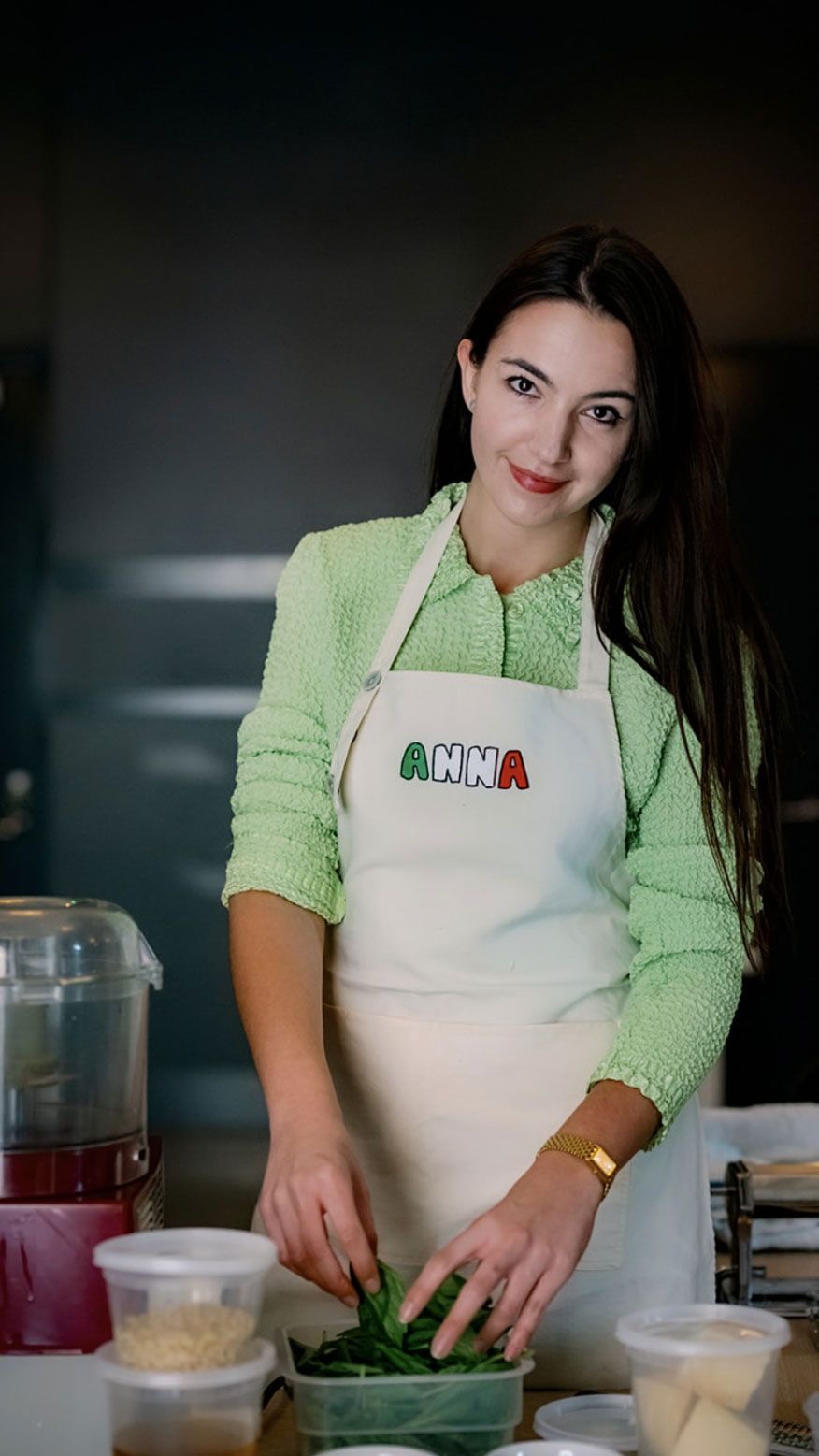 A smiling woman wearing an "ANNA" apron sorts spinach leaves.