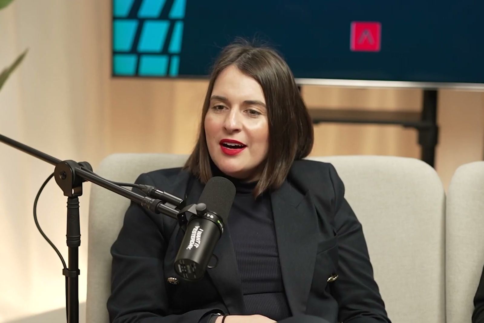 A woman in a black blazer talking at a microphone on a pale sofa