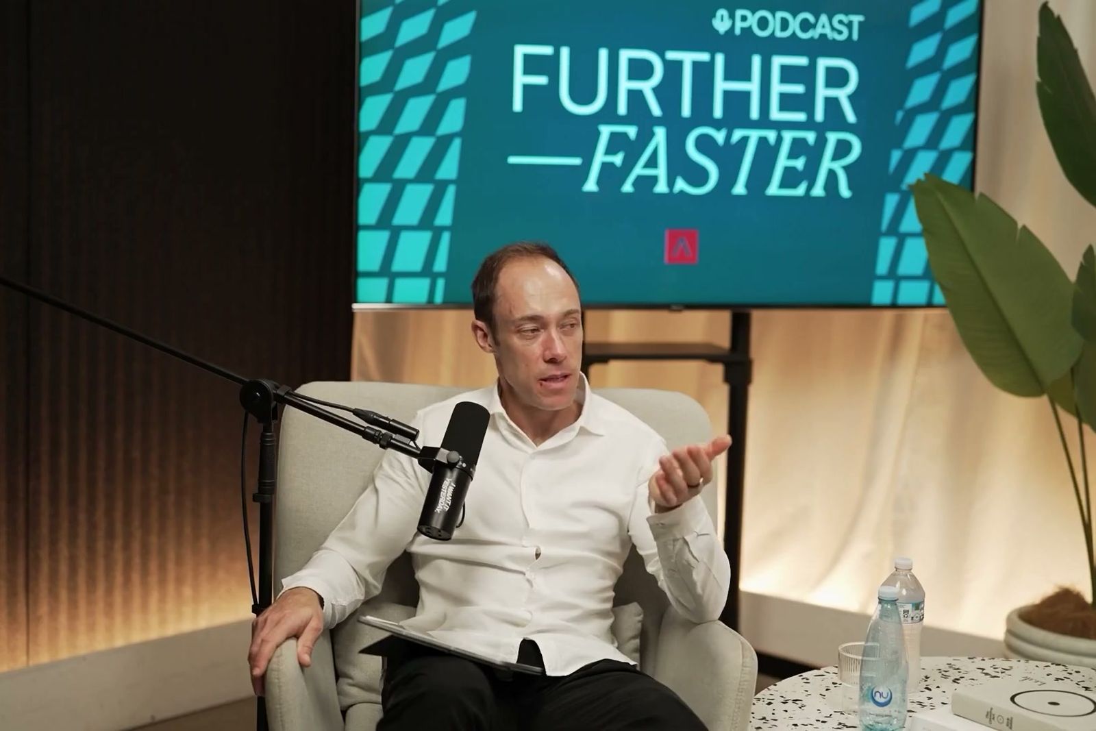 A man in a white shirt gesturing as he talks in an armchair under the Further, Faster sign