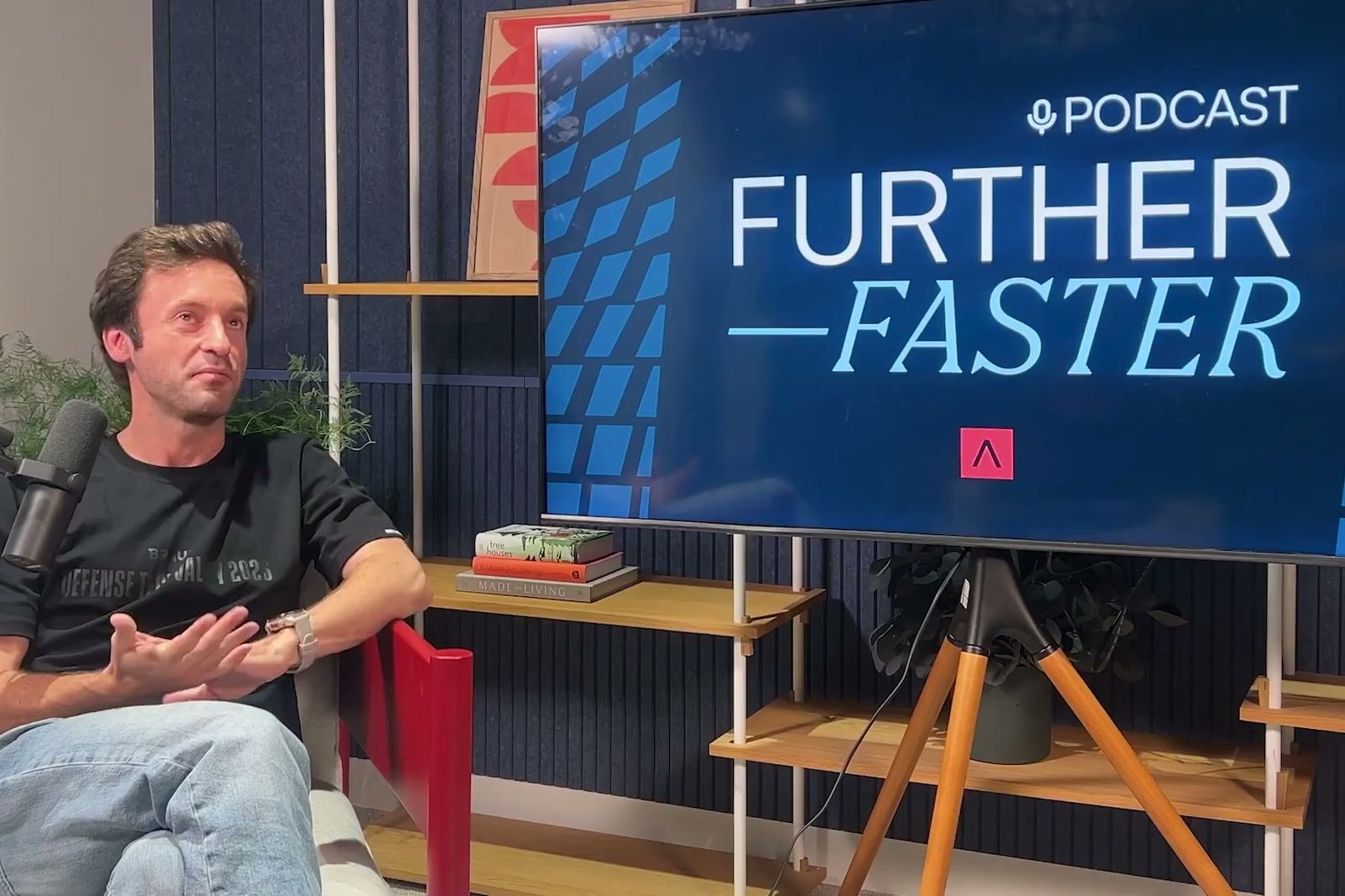 Guiam Wainwright talking with his hands beside a screen showing the Further, Faster logo