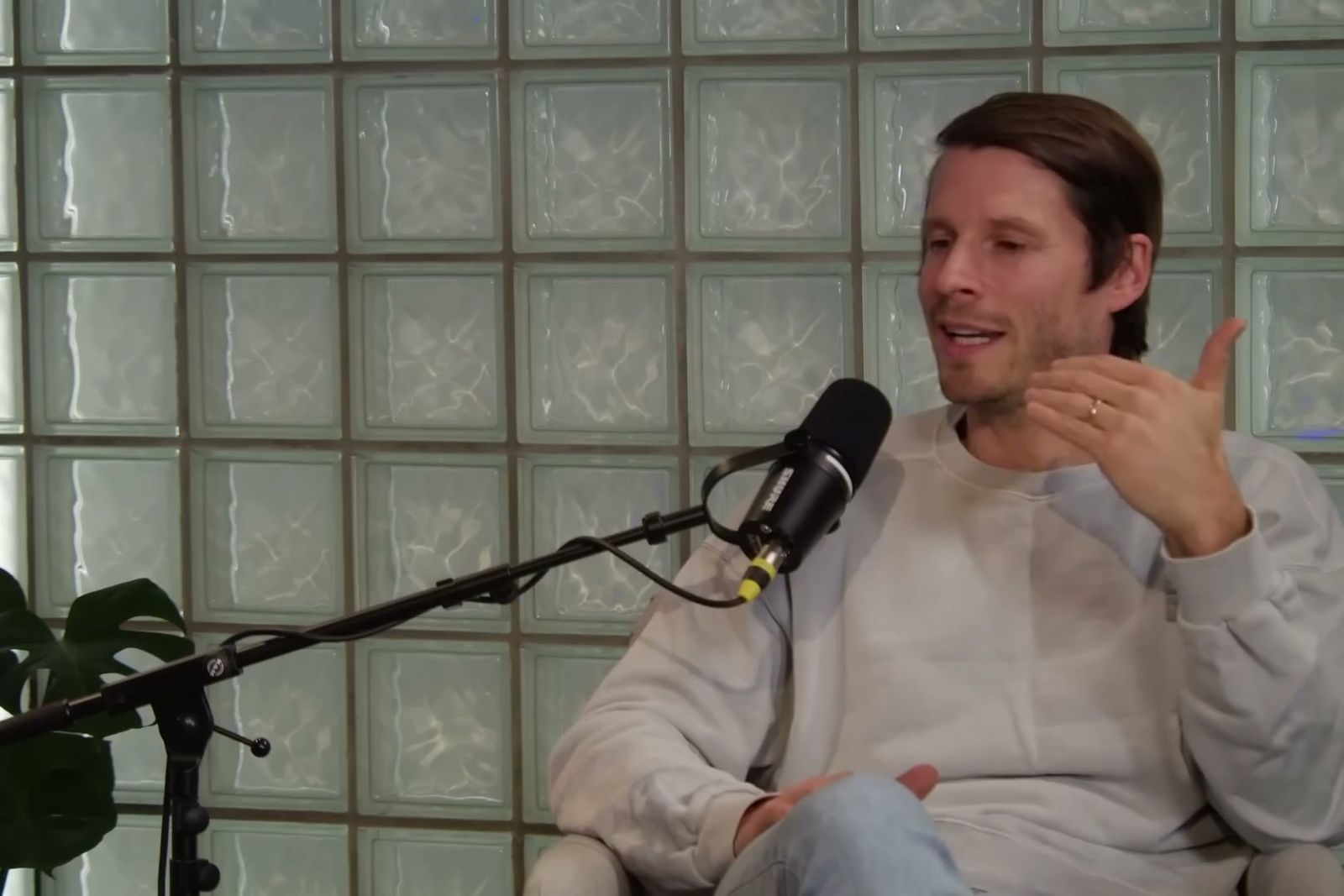 A man in a grey sweatshirt talking with his hands at a microphone in front of a glass-block wall