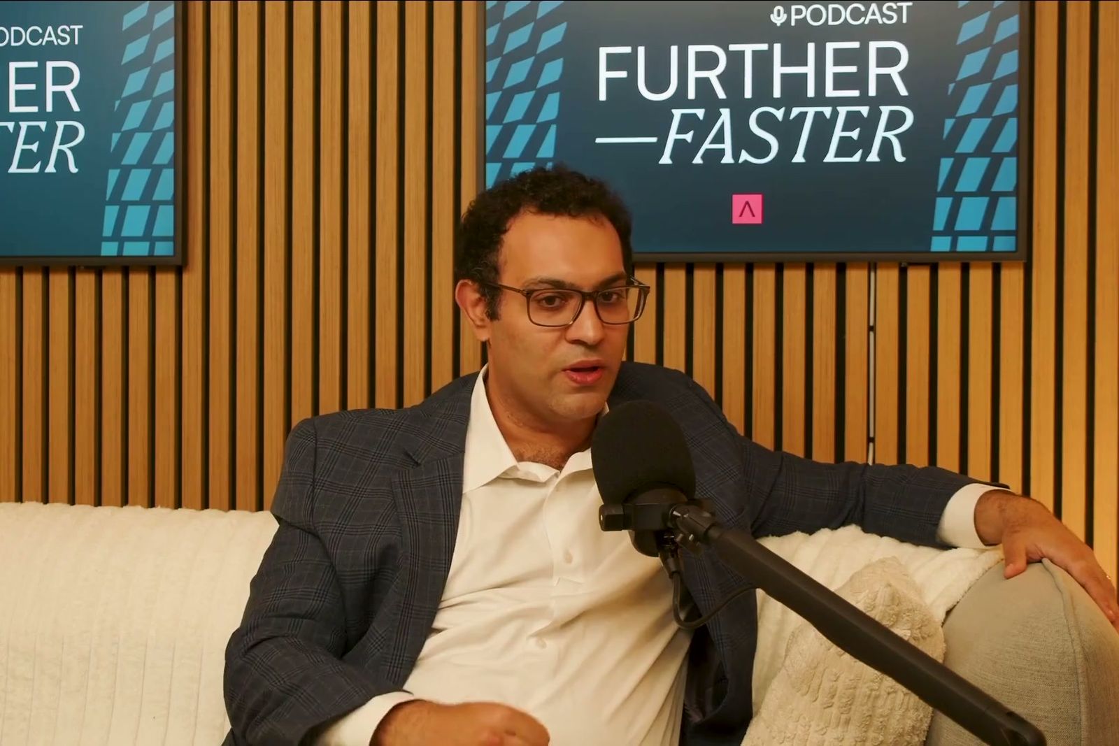 A man in glasses and a blazer talking on a sofa under the Further, Faster sign