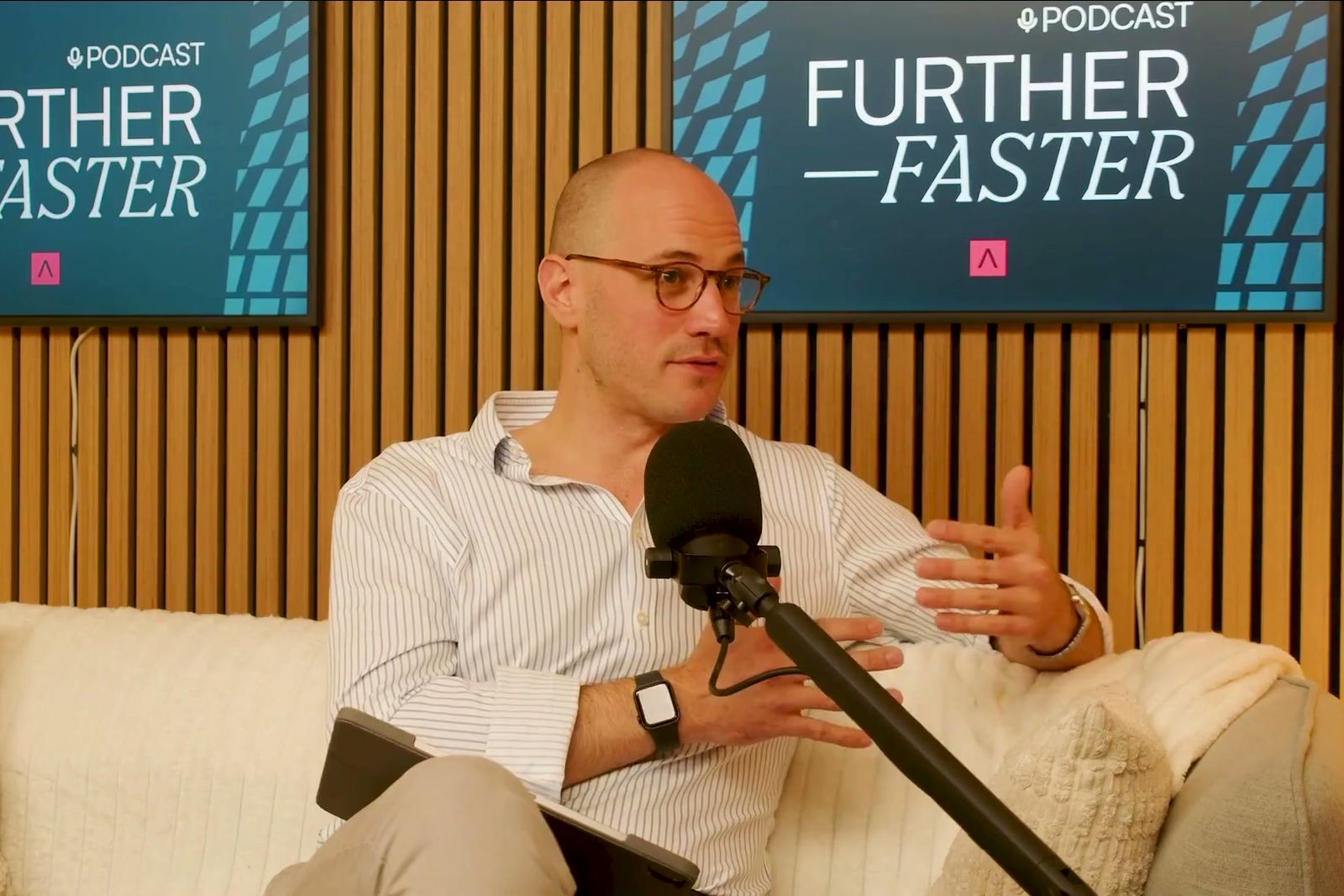 A man in glasses and a striped shirt gesturing as he talks on a sofa under the Further, Faster sign