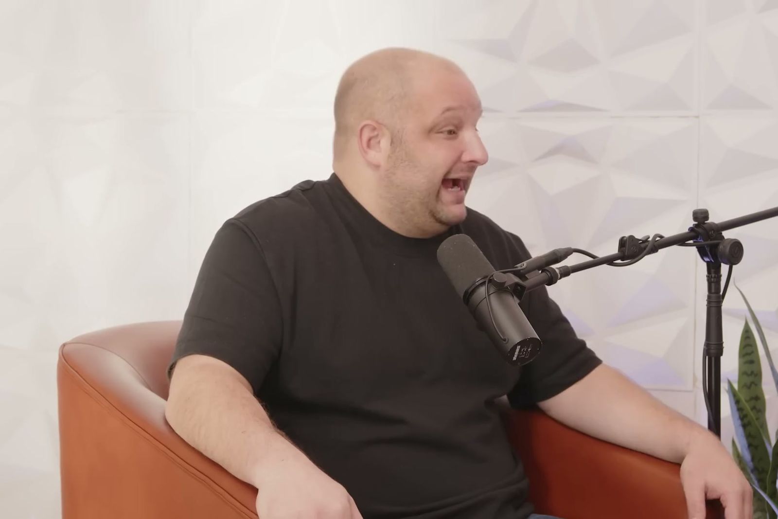 A man in a black T-shirt talking animatedly in a tan leather armchair at a microphone