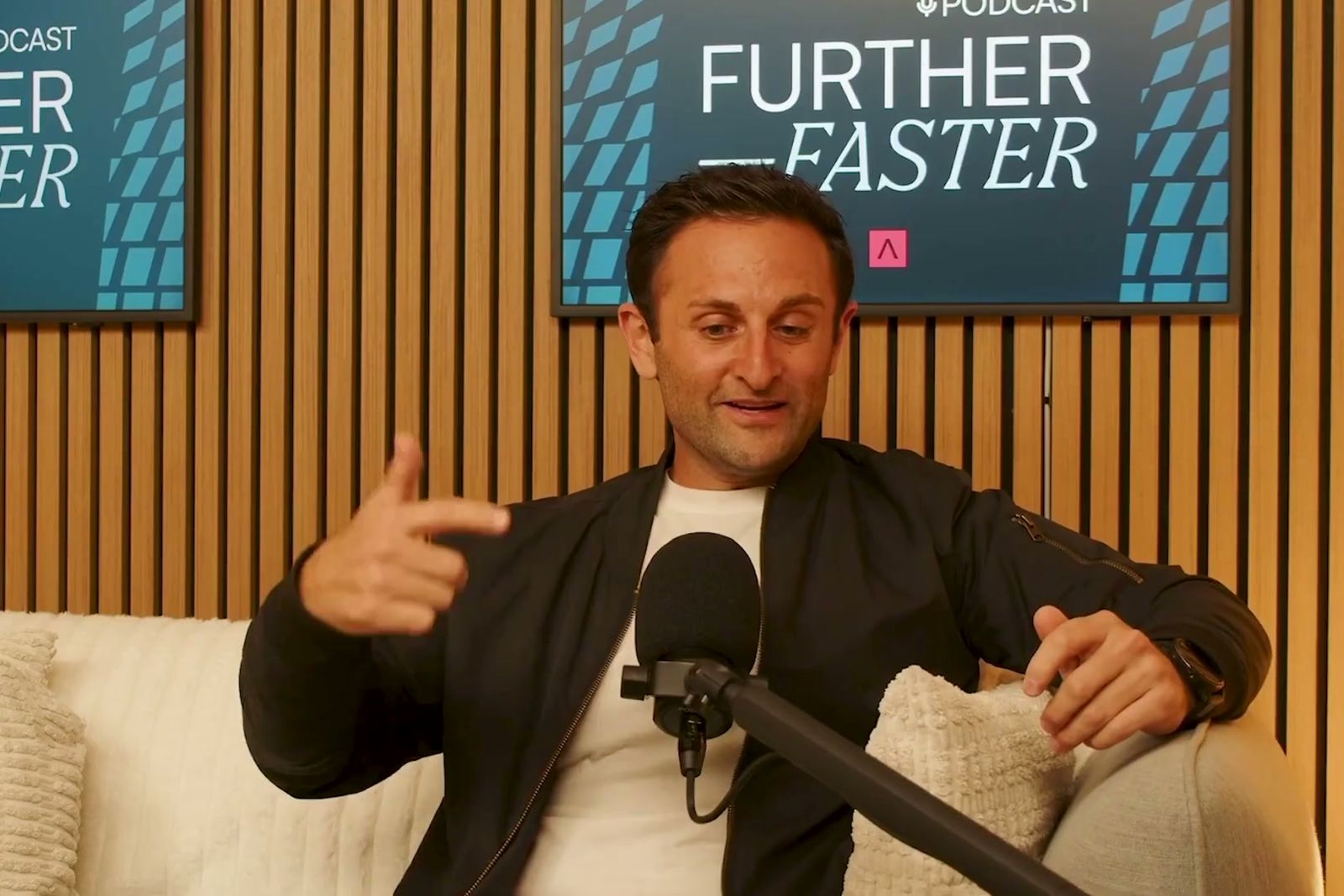 A man in a dark bomber jacket gesturing as he talks on a sofa under the Further, Faster sign