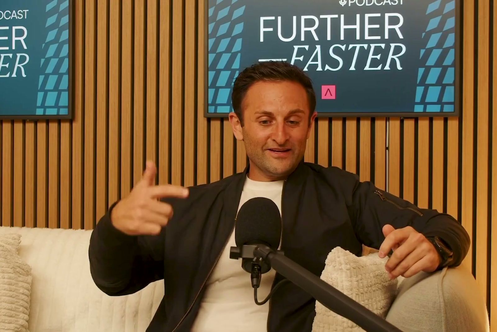 A man in a dark bomber jacket gesturing as he talks on a sofa under the Further, Faster sign