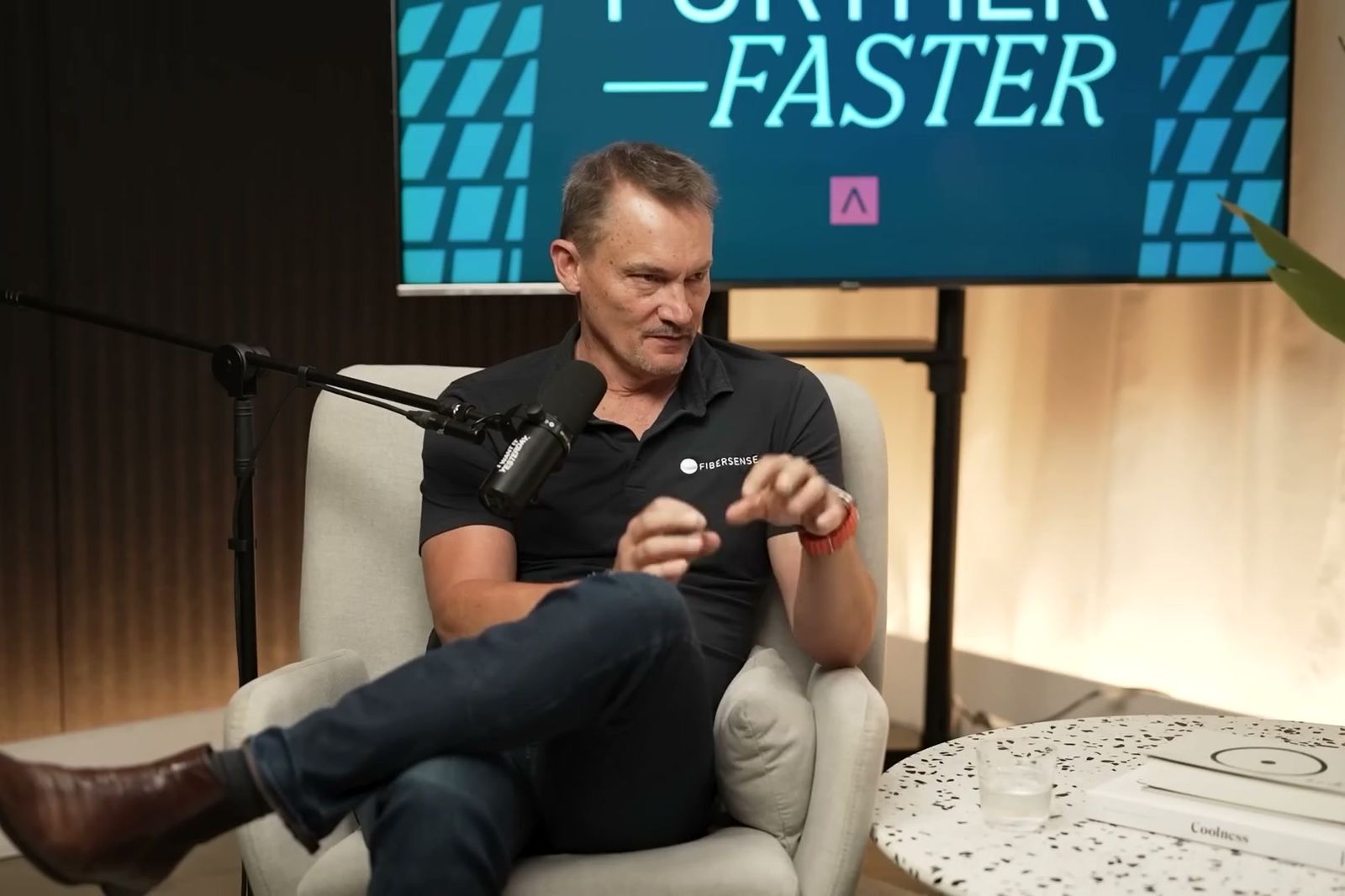 Mark Englund talking with his hands in an armchair under the Further, Faster sign