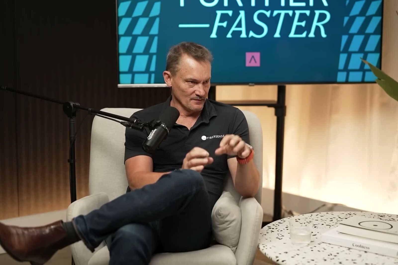 Mark Englund talking with his hands in an armchair under the Further, Faster sign