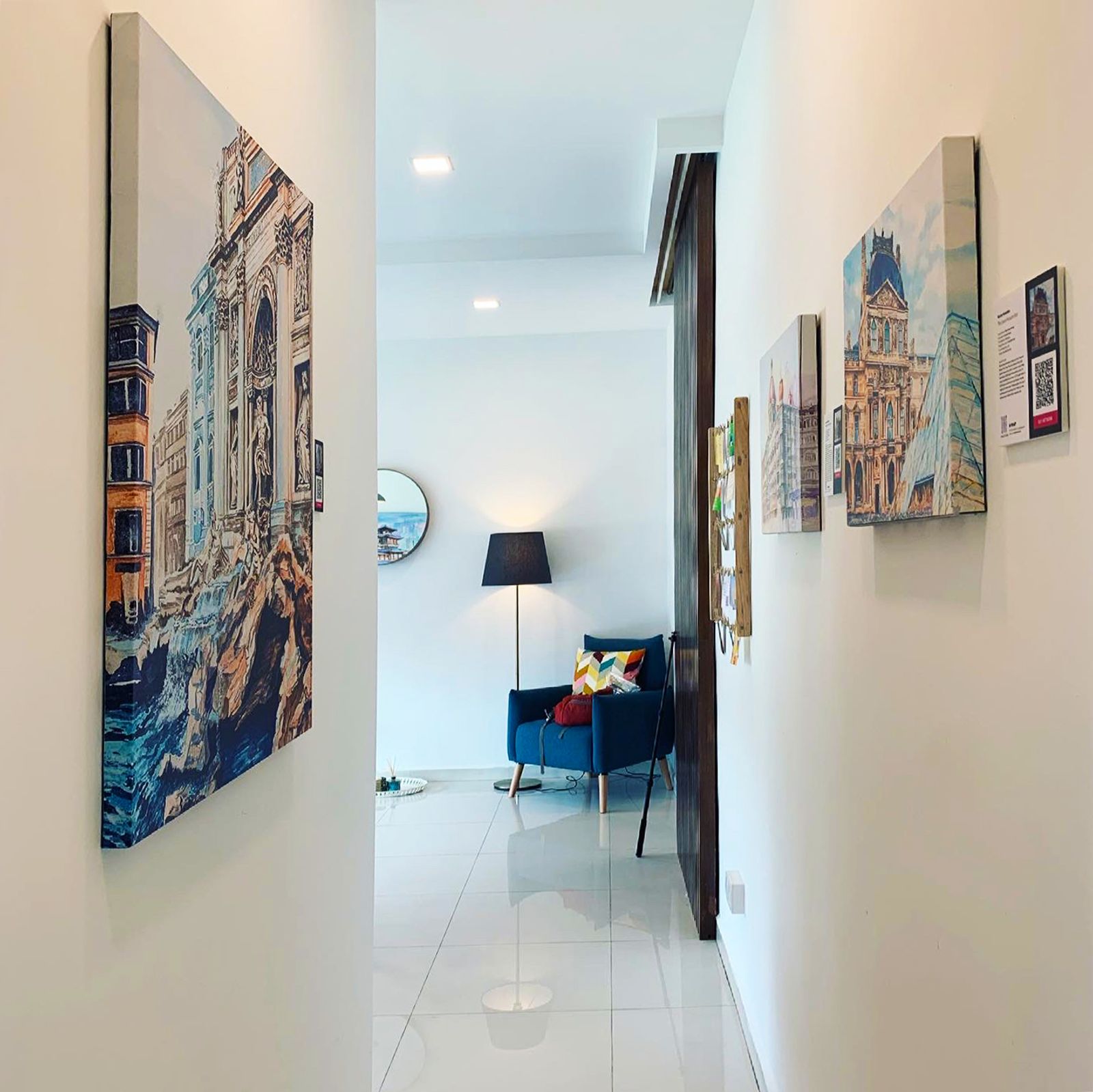 Image of hallway with artwork hanging on the walls looking through to a room with chair and lamp 