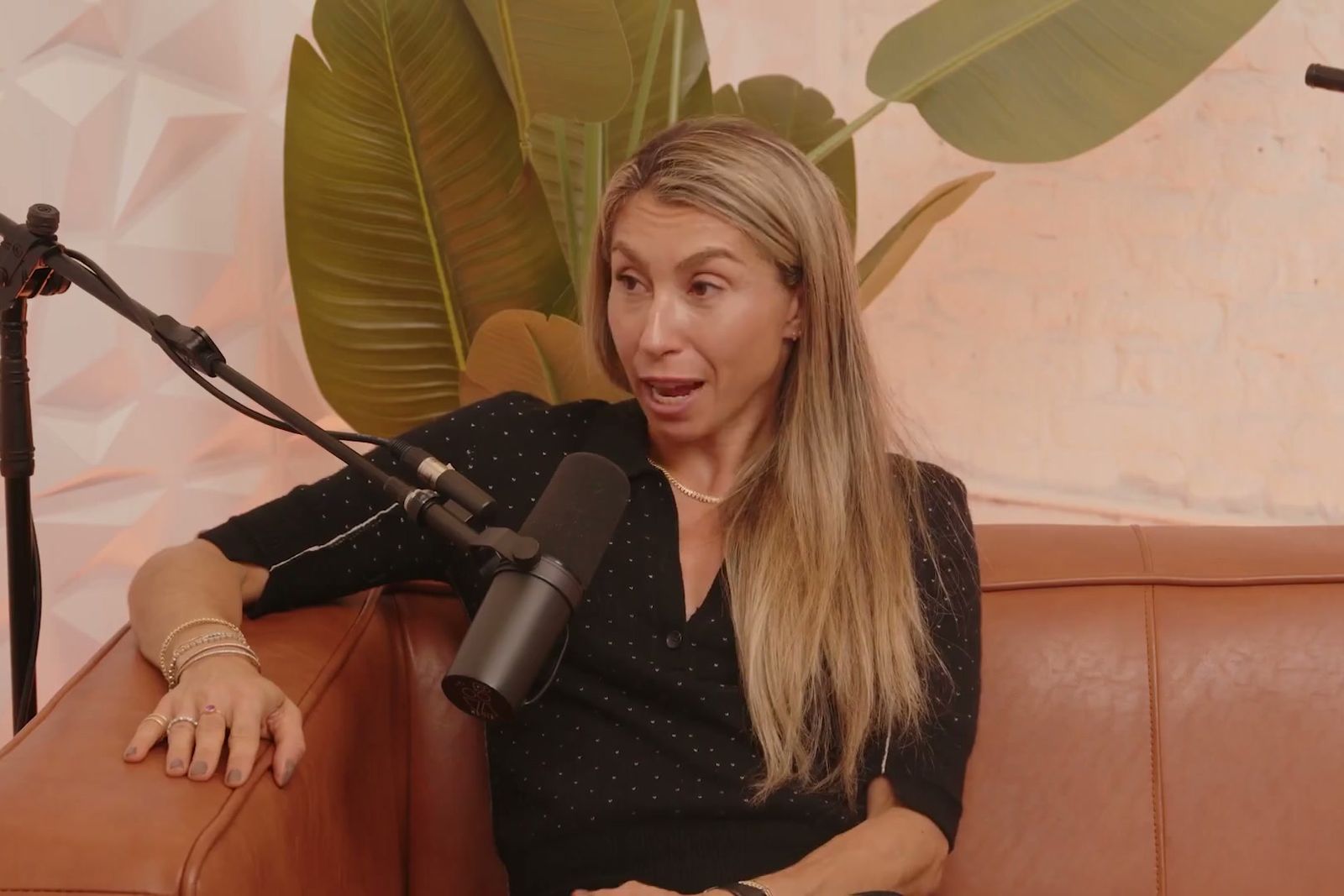 Jenny Fleiss talking at a microphone on a tan leather sofa in front of a large leafy plant
