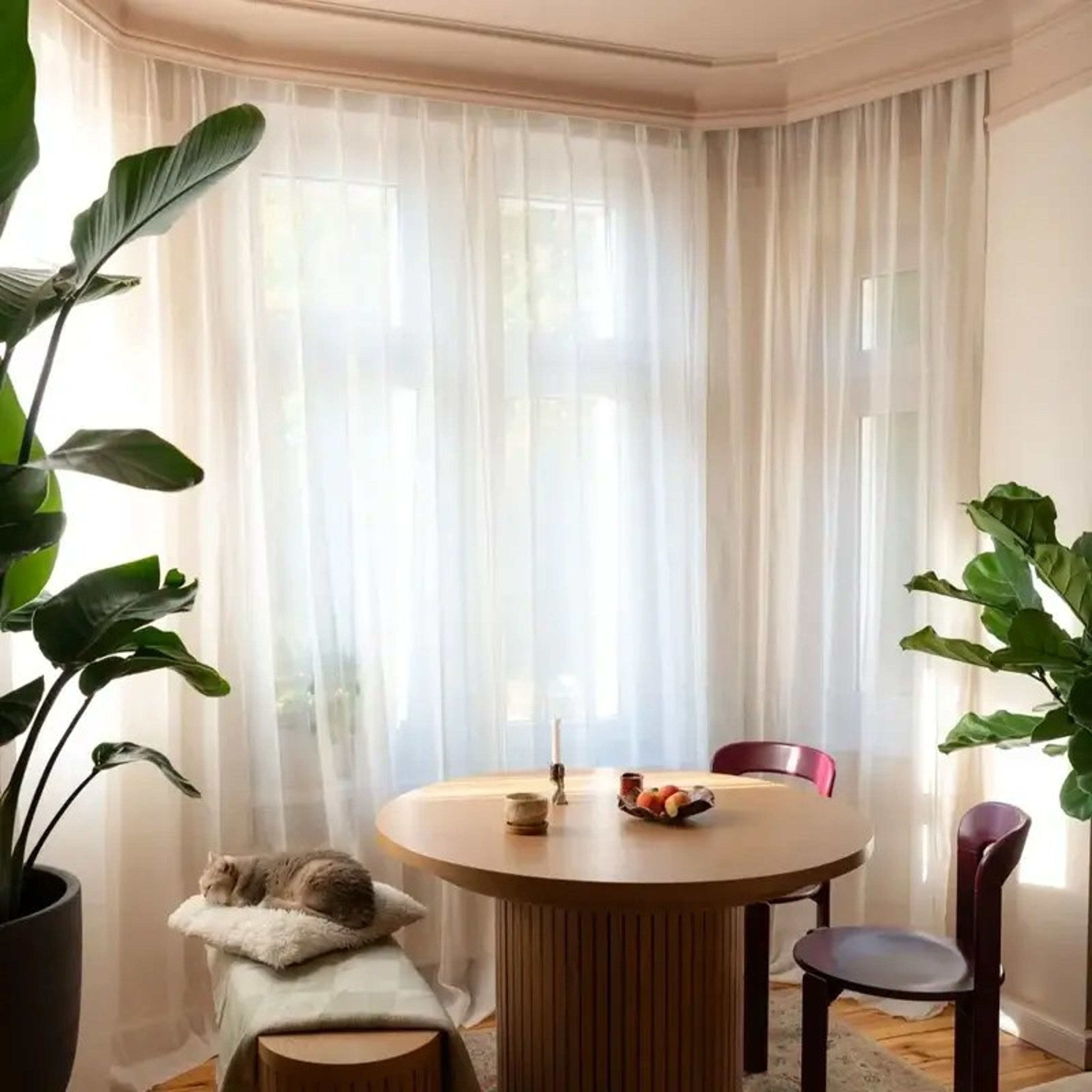 Bay window dining nook with plain white sheer curtains, a round wooden table and burgundy chairs