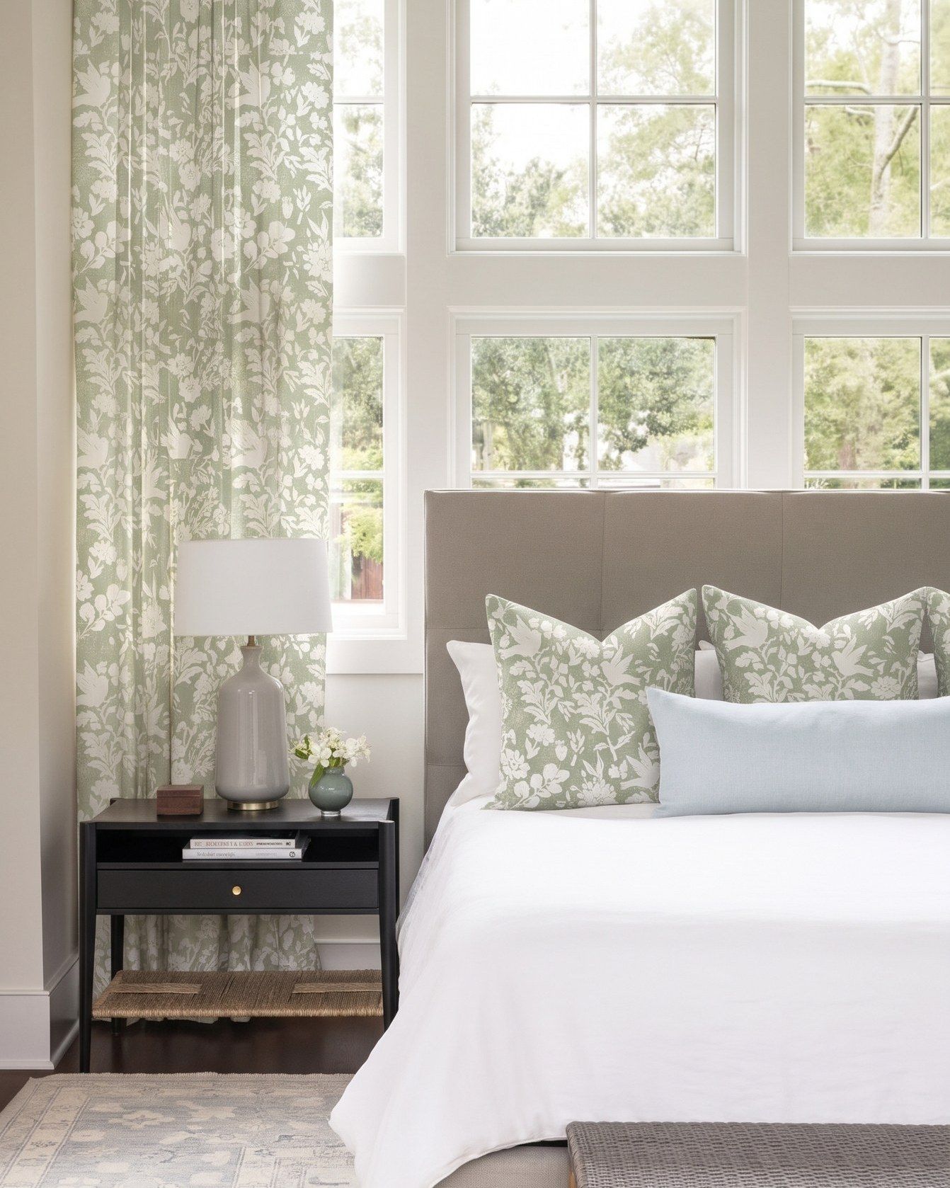 Sage green and white floral curtains beside a bed with matching floral cushions, a pale blue pillow and a black nightstand, in a bedroom with tall white windows