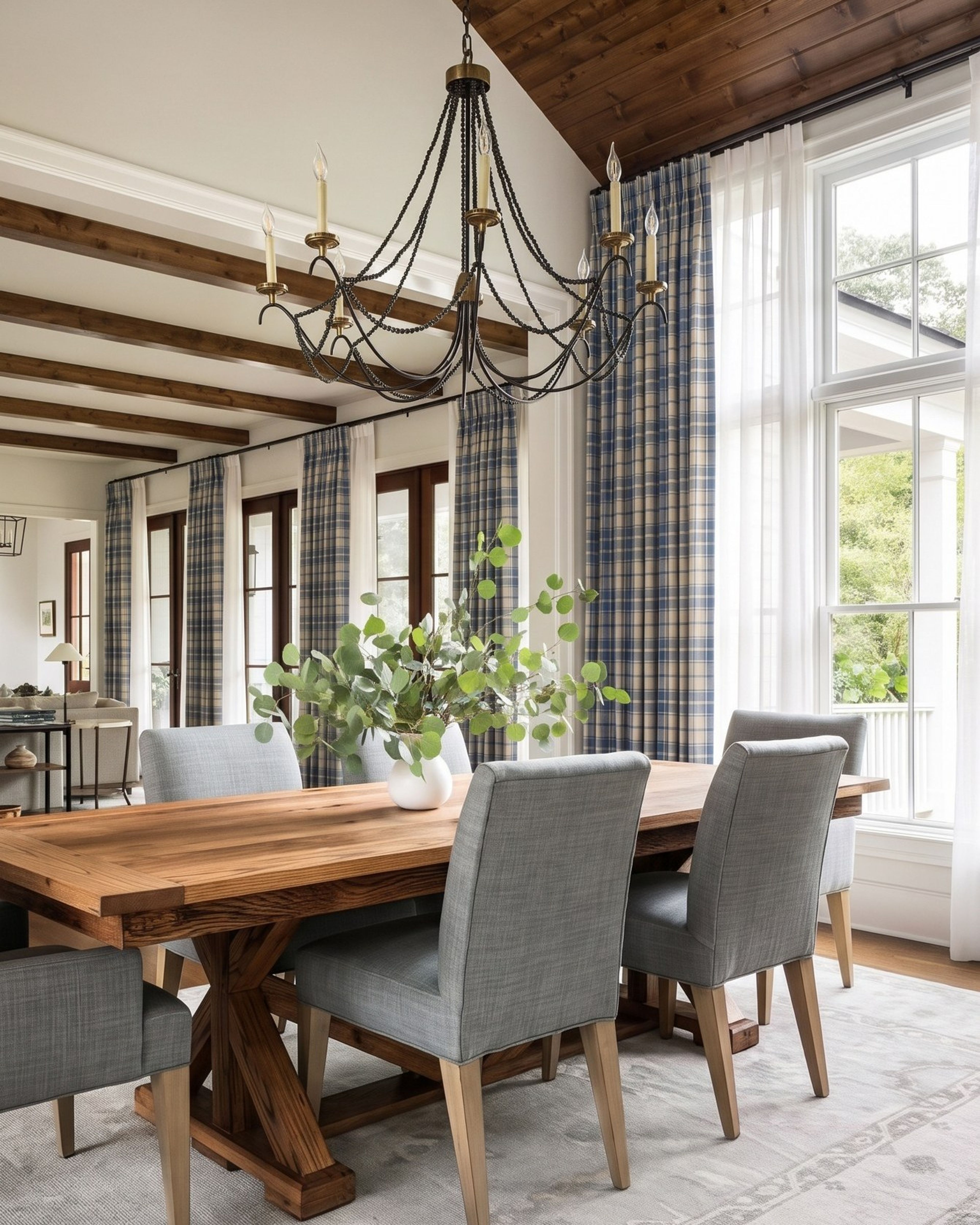 A dining room with blue and cream plaid curtains layered over white sheers, a rustic wooden table, gray upholstered chairs and an iron chandelier